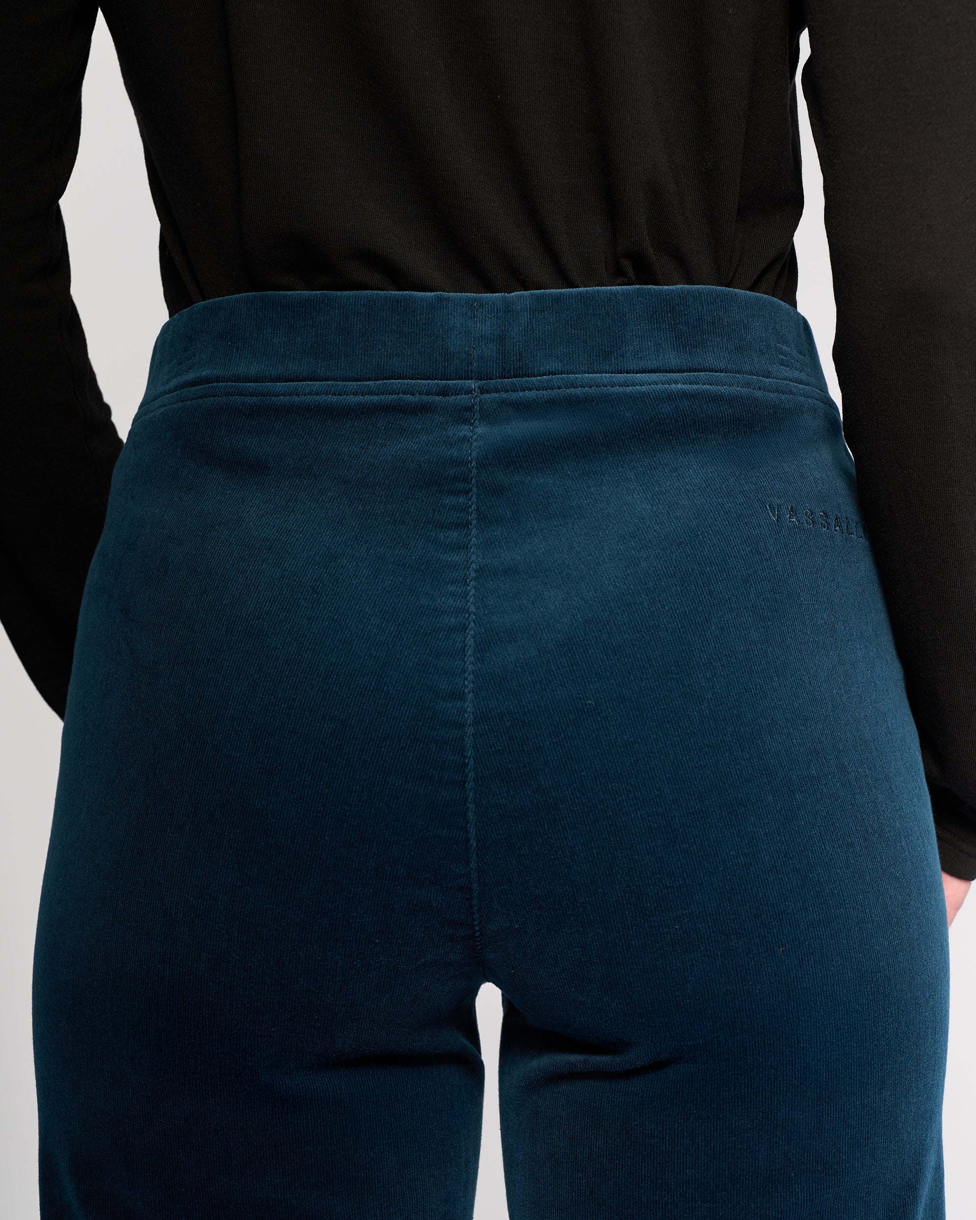 A person wearing a black long-sleeve top and Vassalli Loft Cord Pull On pants in Petrol, shown from the back at waist height, with subtle branding visible near the pull-on waist.