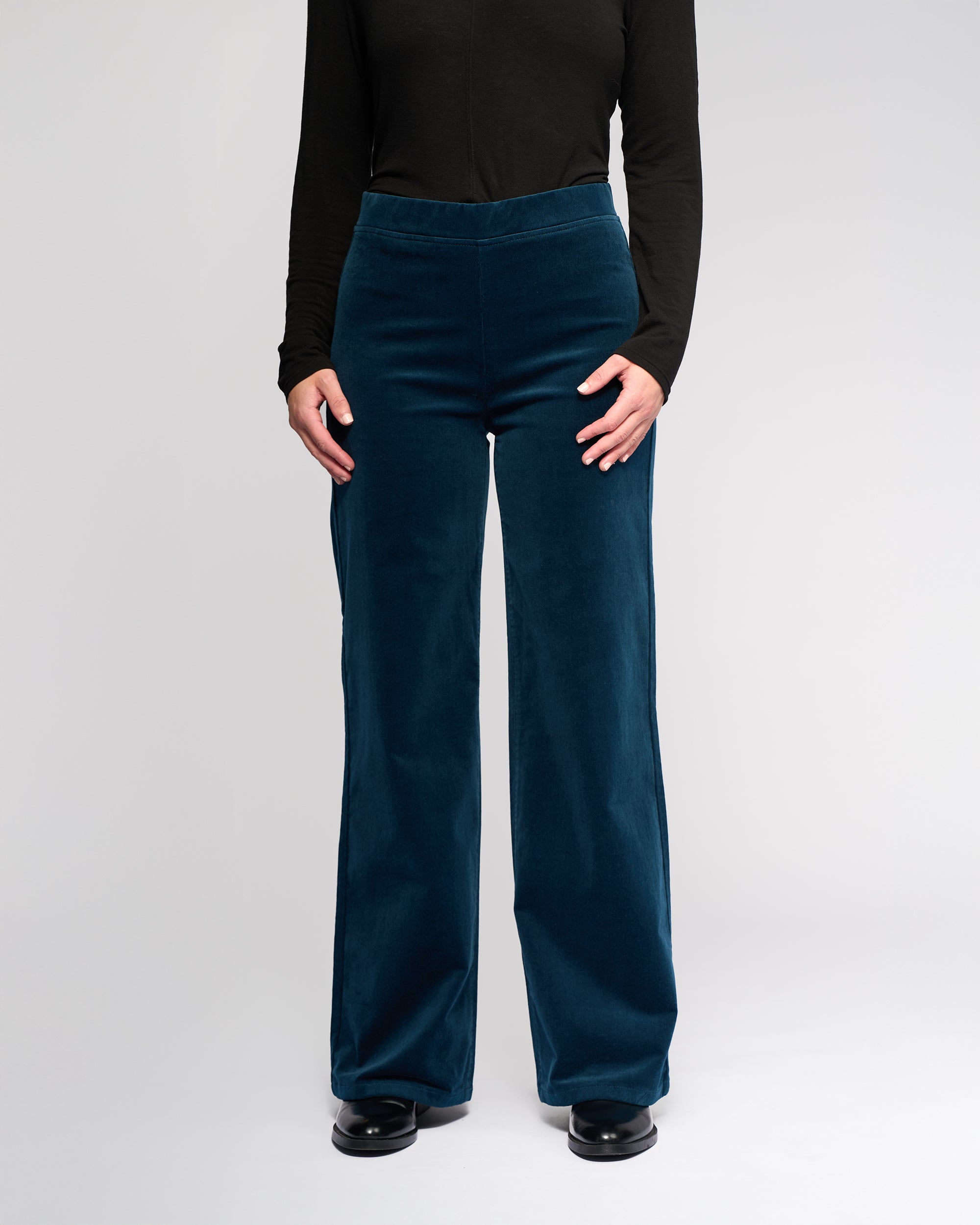 Wearing a black long-sleeve shirt and Vassalli Loft Cord Pull On - Petrol wide-leg pants with a comfy pull-on waist, paired with black shoes, the person stands against a plain light background, hands at their sides.