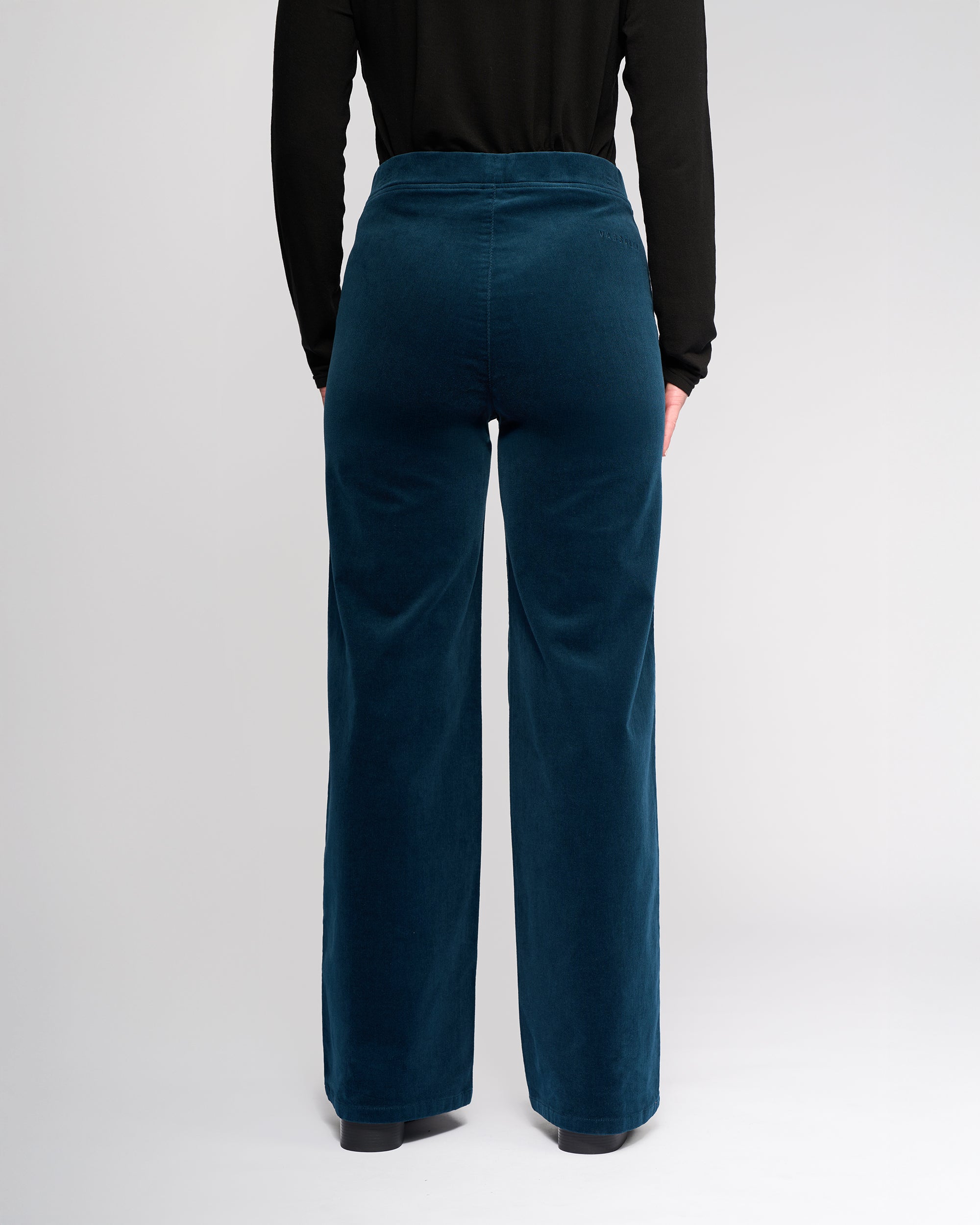 A person wearing a long-sleeve black top and Vassalli Loft Cord Pull On - Petrol high-waisted, wide-leg pants with a stretch pull-on waist stands facing away from the camera against a plain light background.