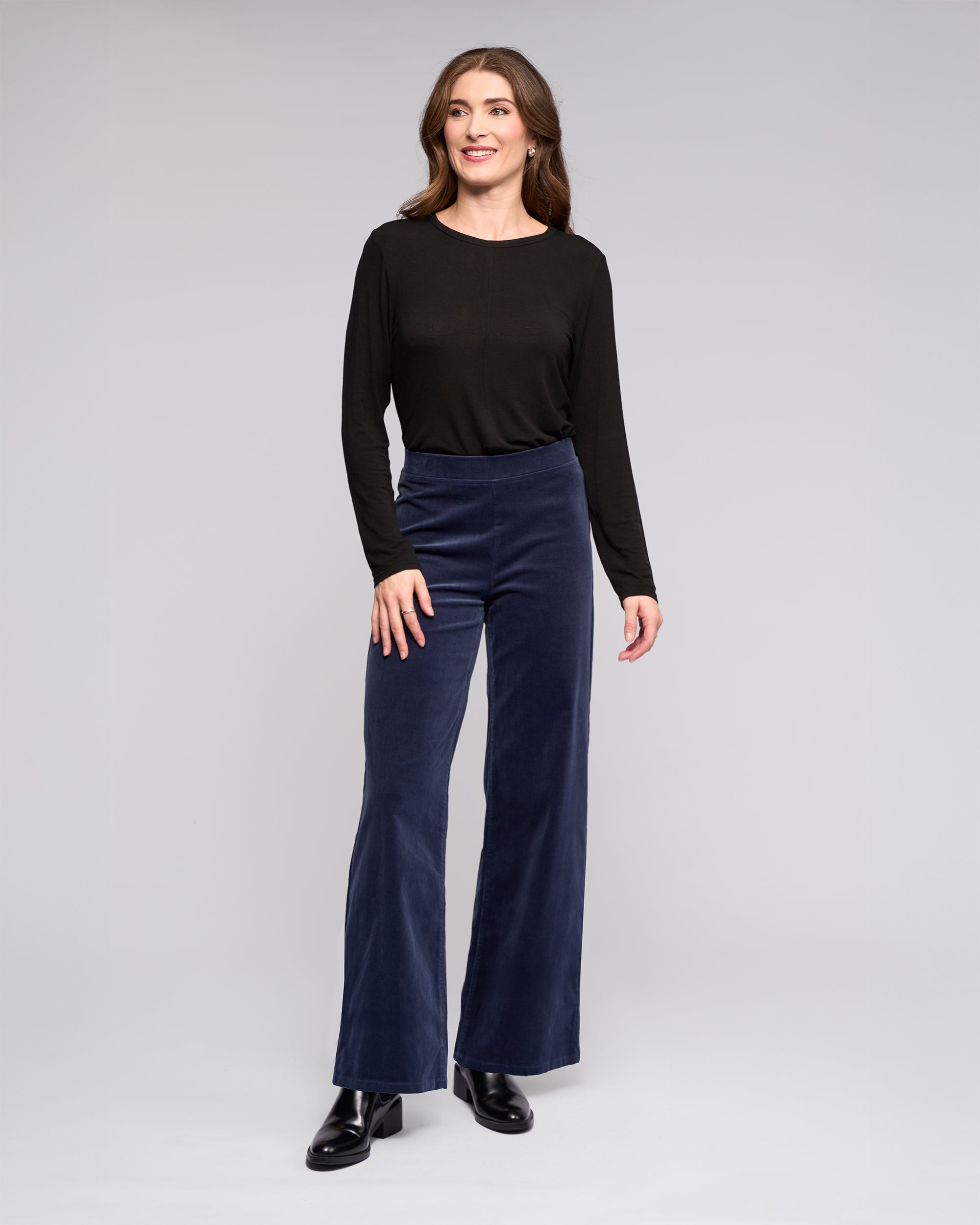 A person with long wavy brown hair smiles slightly while looking off to the side, wearing a black long-sleeve top, black boots, and Vassalli's Loft Cord Pull On - Slate high-waisted wide-leg pants against a plain gray background.