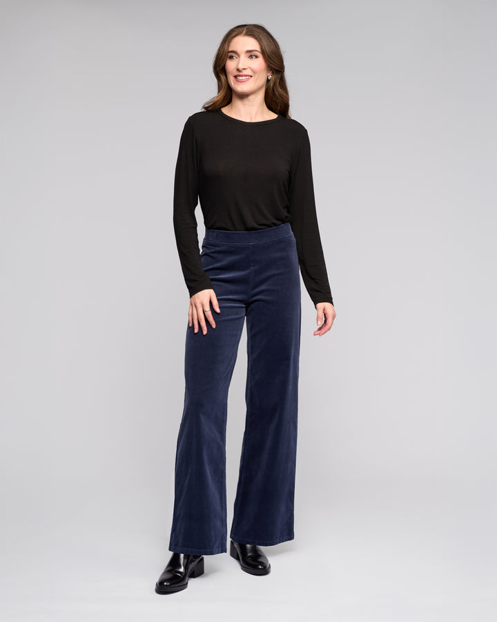 A person with long wavy brown hair smiles slightly while looking off to the side, wearing a black long-sleeve top, black boots, and Vassalli's Loft Cord Pull On - Slate high-waisted wide-leg pants against a plain gray background.