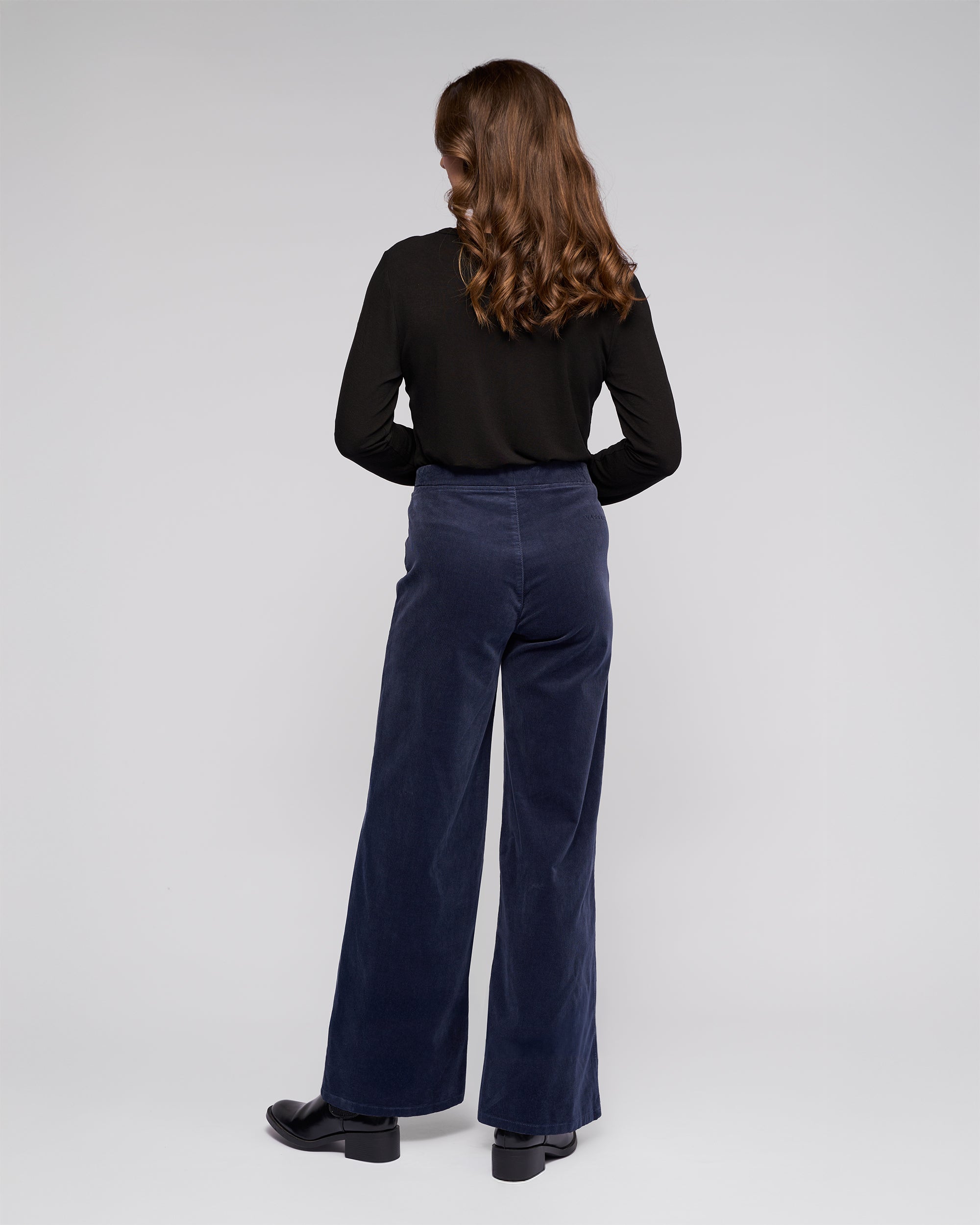 A person with long, wavy brown hair stands facing away, wearing a black long-sleeve top, Vassalli Loft Cord Pull On pants in Slate, and black shoes against a plain light gray background.