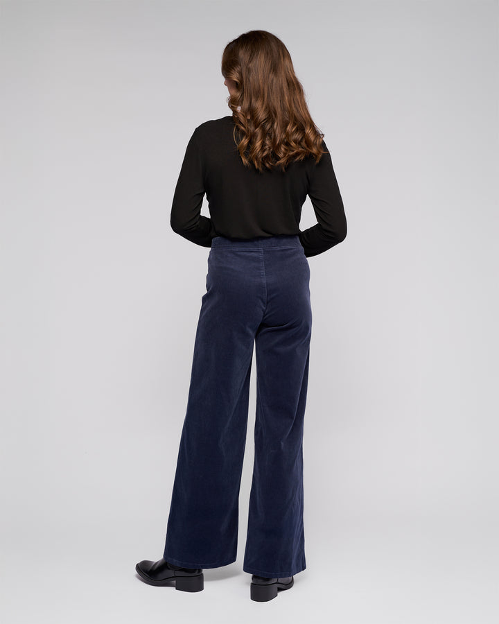 A person with long, wavy brown hair stands facing away, wearing a black long-sleeve top, Vassalli Loft Cord Pull On pants in Slate, and black shoes against a plain light gray background.