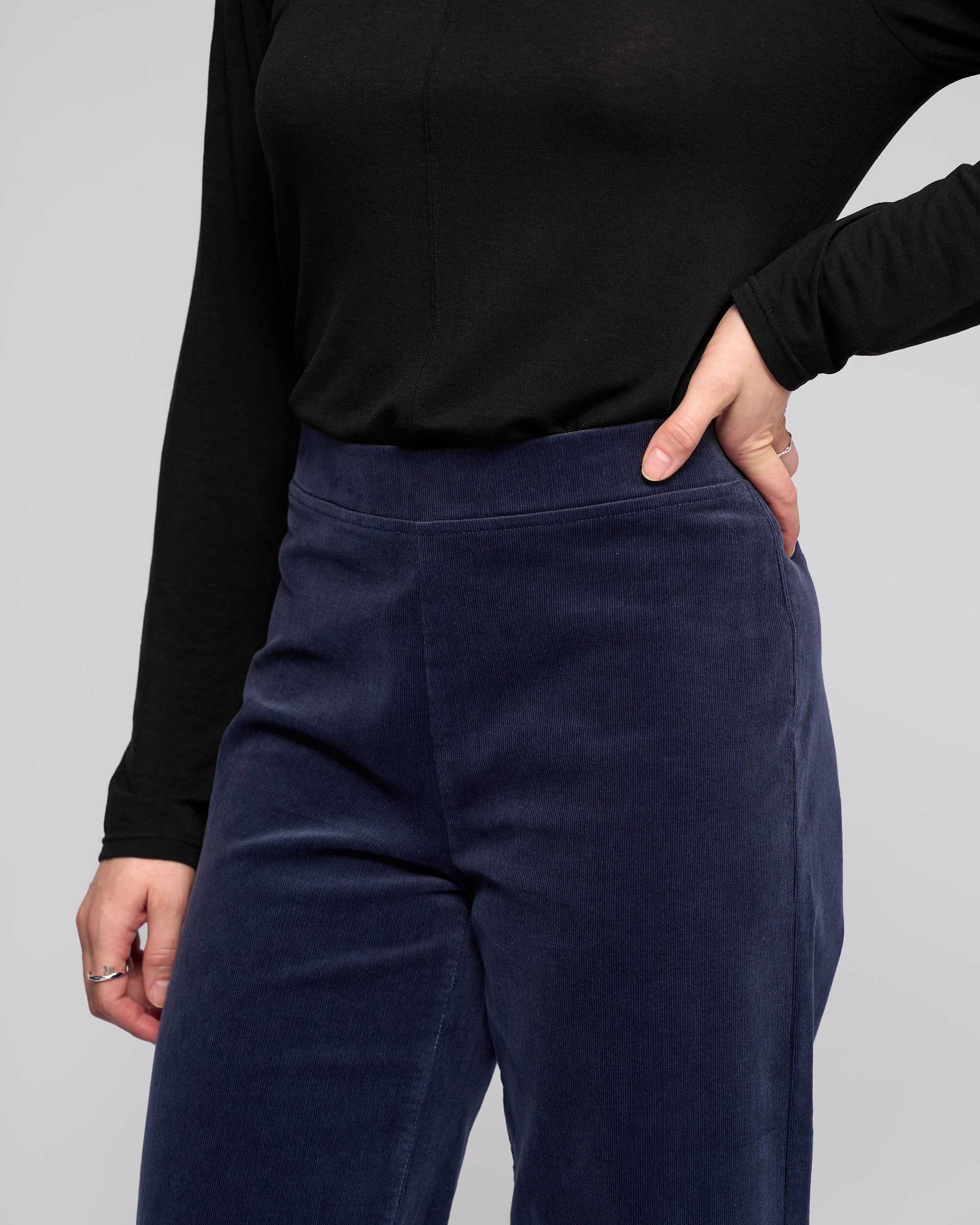 A person stands against a plain light background wearing Vassalli's Loft Cord Pull On pants in Slate, featuring a high-waisted, wide-leg style with an elastic waistband and paired with a long-sleeved black top.