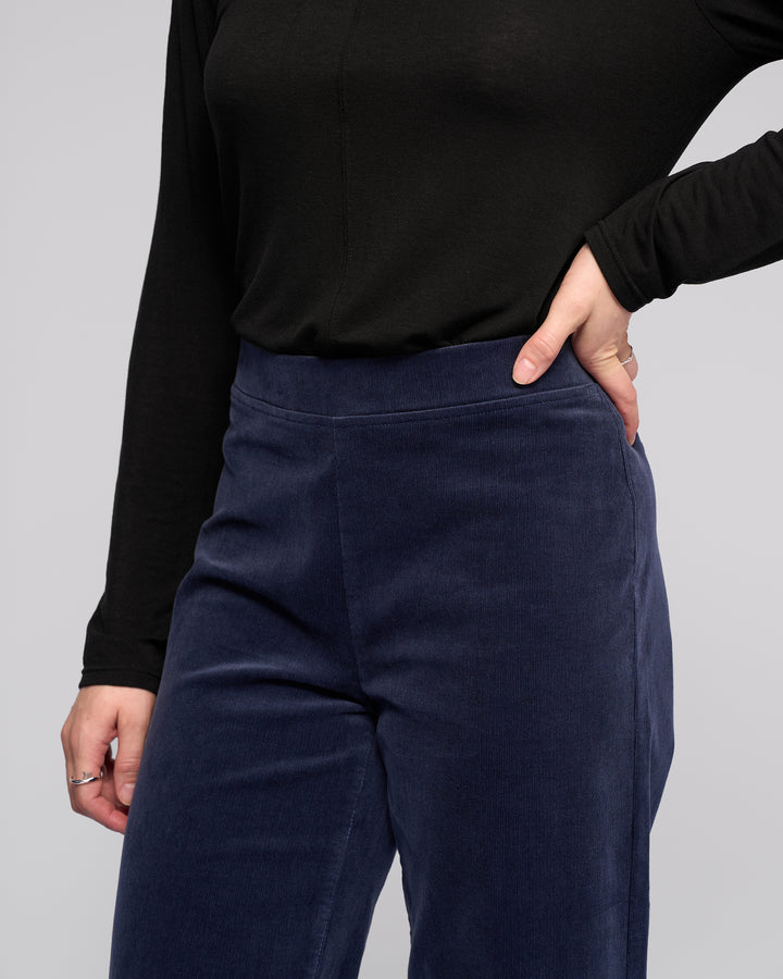 A person stands against a plain light background wearing Vassalli's Loft Cord Pull On pants in Slate, featuring a high-waisted, wide-leg style with an elastic waistband and paired with a long-sleeved black top.