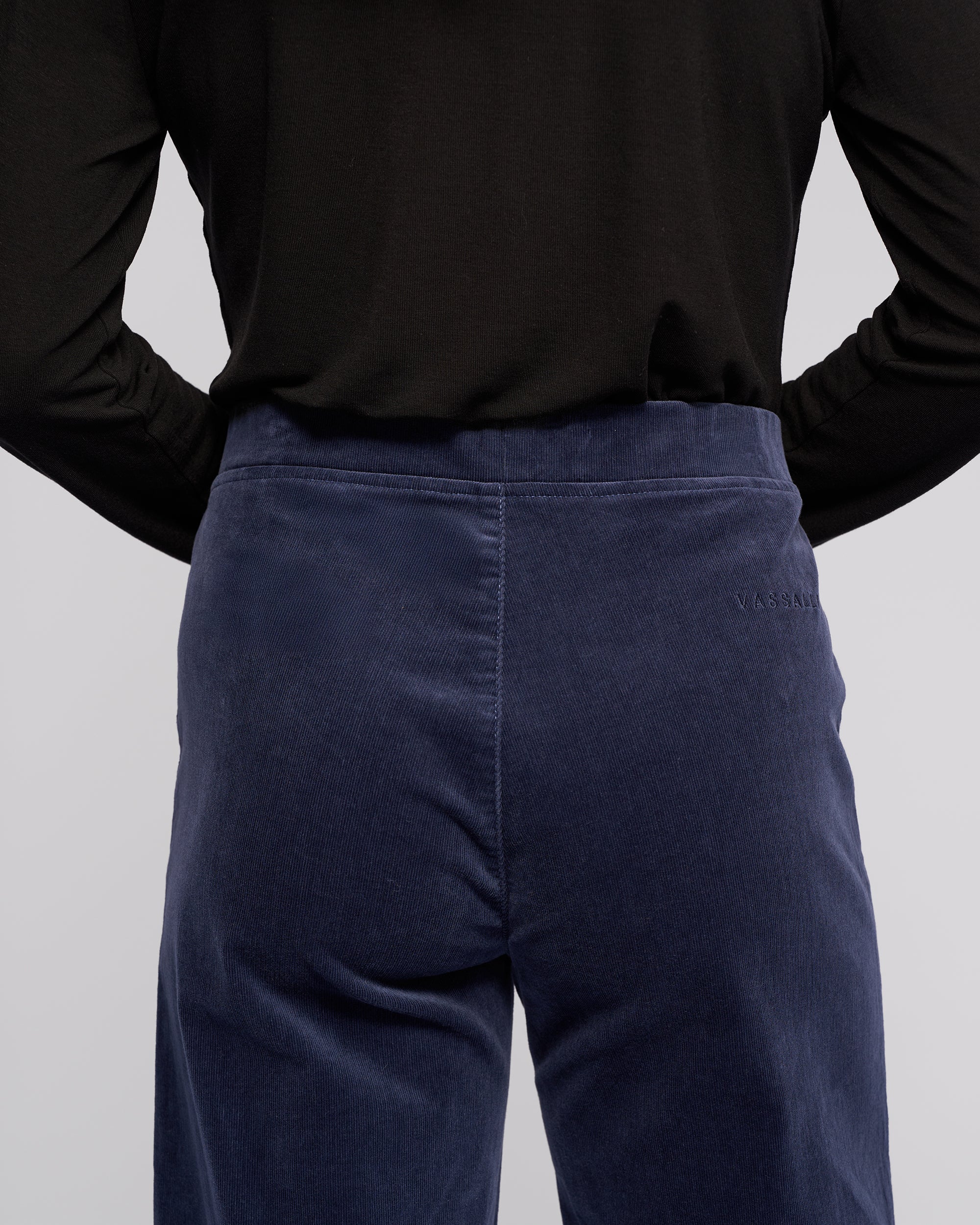 A person is shown from the back wearing a black long-sleeve top and high-waisted, dark blue Loft Cord Pull On - Slate pants by Vassalli, which feature subtle stitching details and the brand name embroidered on the right side.