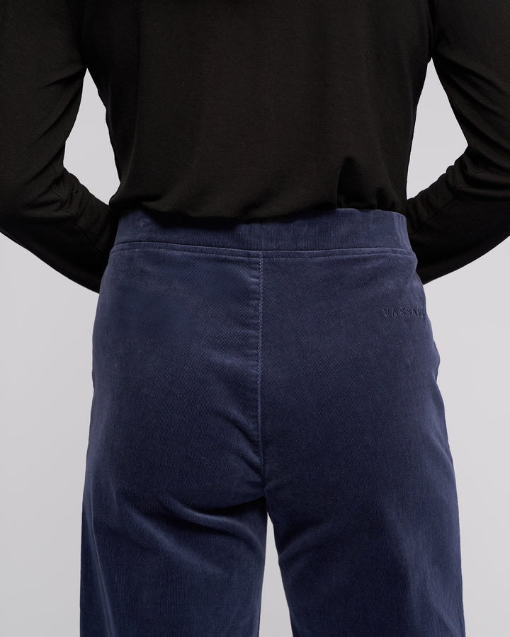 A person is shown from the back wearing a black long-sleeve top and high-waisted, dark blue Loft Cord Pull On - Slate pants by Vassalli, which feature subtle stitching details and the brand name embroidered on the right side.