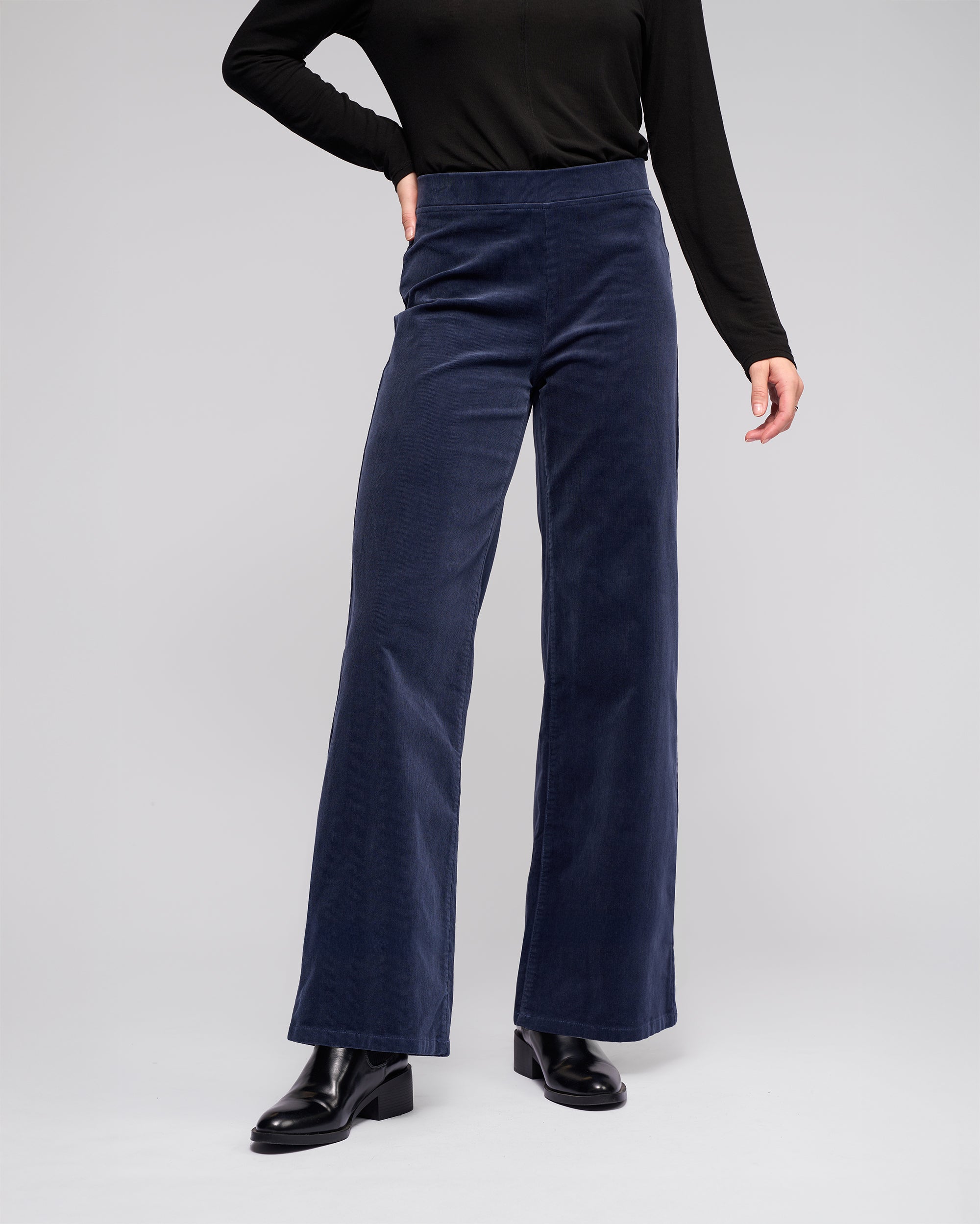 A person wearing Vassalli Loft Cord Pull On - Slate pants with a black long-sleeve top and black ankle boots stands against a plain light gray background.