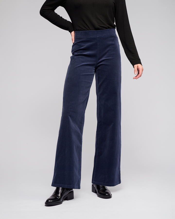 A person wearing Vassalli Loft Cord Pull On - Slate pants with a black long-sleeve top and black ankle boots stands against a plain light gray background.