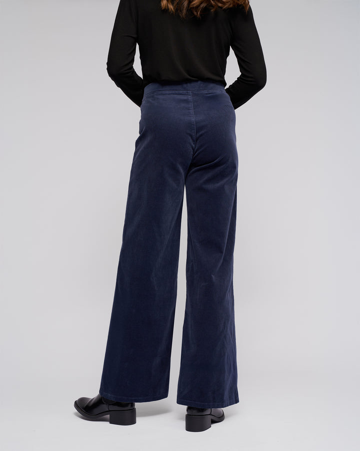 A person stands facing away from the camera, wearing Vassalli's Loft Cord Pull On - Slate pants with an elastic waistband, a black long-sleeve top, and black shoes against a plain light gray background.