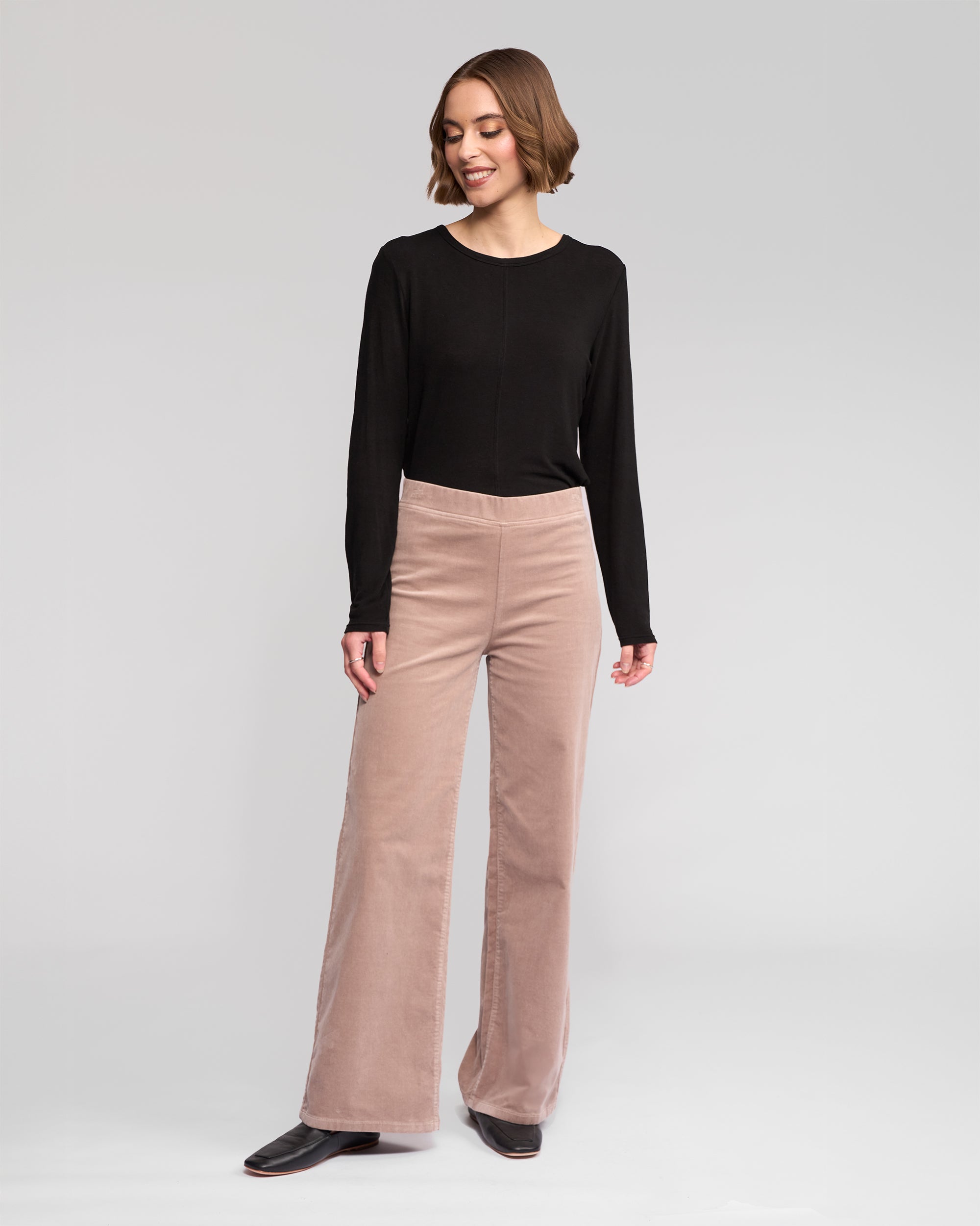 A woman stands against a plain background wearing a black long-sleeve top, Vassalli Loft Cord Pull On - Twig pants in a comfortable style, and black shoes, looking slightly to the side and smiling.