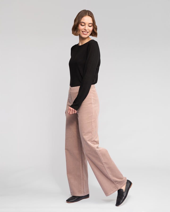 A woman with short brown hair smiles while posing in a black long-sleeve top, Vassalli Loft Cord Pull On - Twig pants, and black shoes against a plain light grey background.