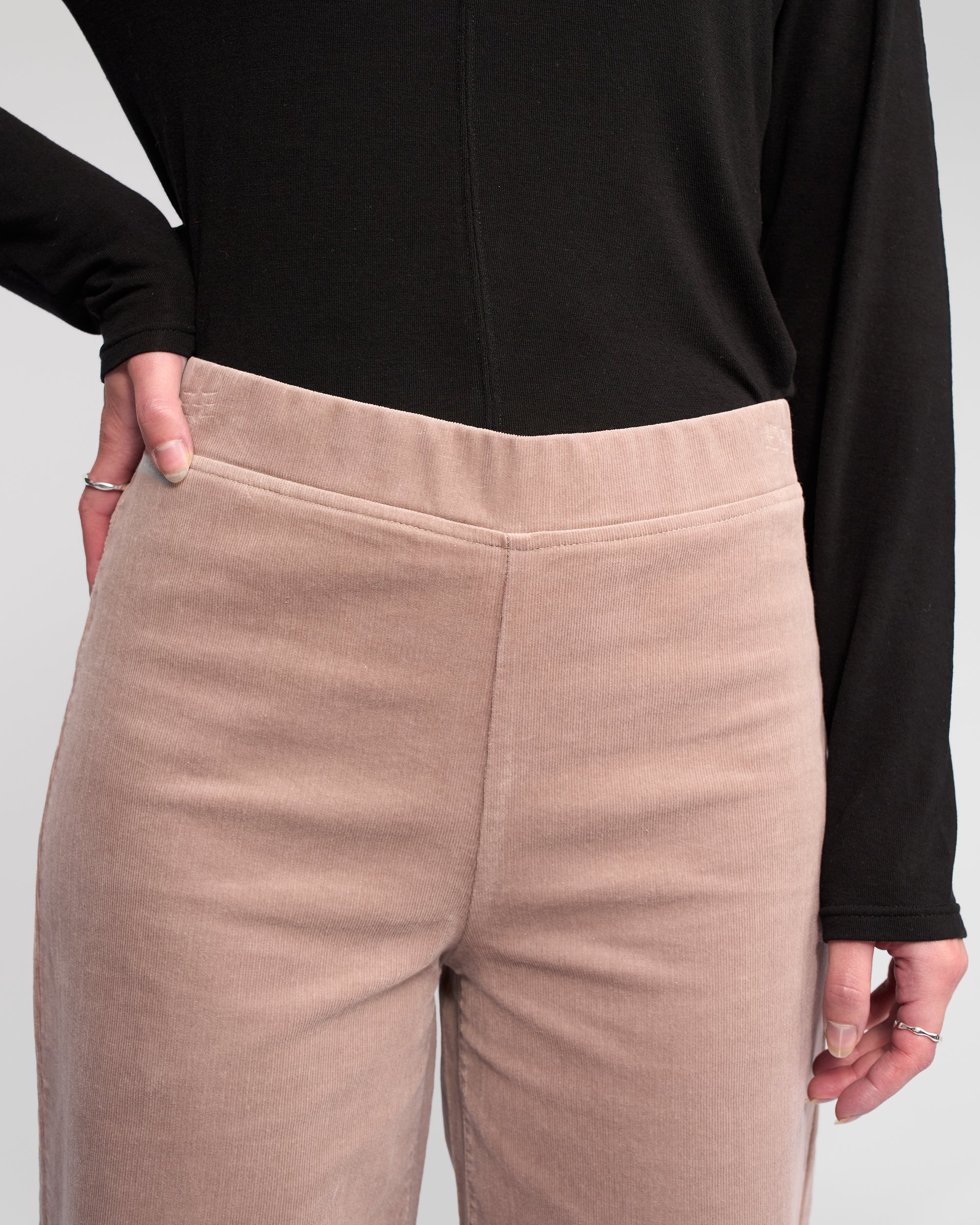 Wearing Vassalli's Loft Cord Pull On - Twig, a person stands with one hand on their hip in a black long-sleeve shirt; only the torso and upper legs are visible.
