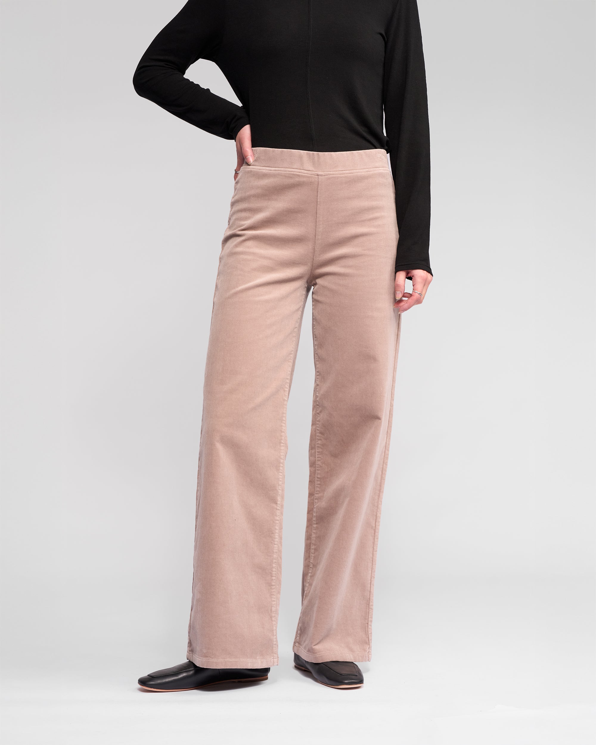 A person wears the Vassalli Loft Cord Pull On - Twig pants with a black long-sleeve top and black shoes, standing with one hand on their hip against a plain light background.
