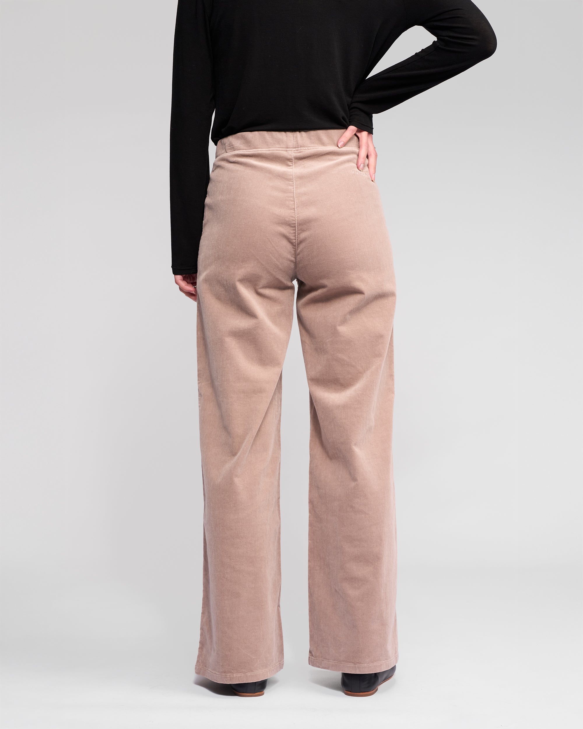A person stands with their back to the camera, wearing a long-sleeve black top and Vassalli Loft Cord Pull On - Twig high-waisted, wide-leg stretch pinwale cord pants, one hand on their hip, against a plain light background.