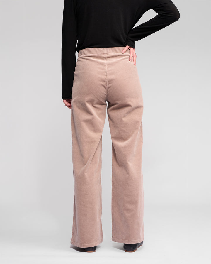A person stands with their back to the camera, wearing a long-sleeve black top and Vassalli Loft Cord Pull On - Twig high-waisted, wide-leg stretch pinwale cord pants, one hand on their hip, against a plain light background.