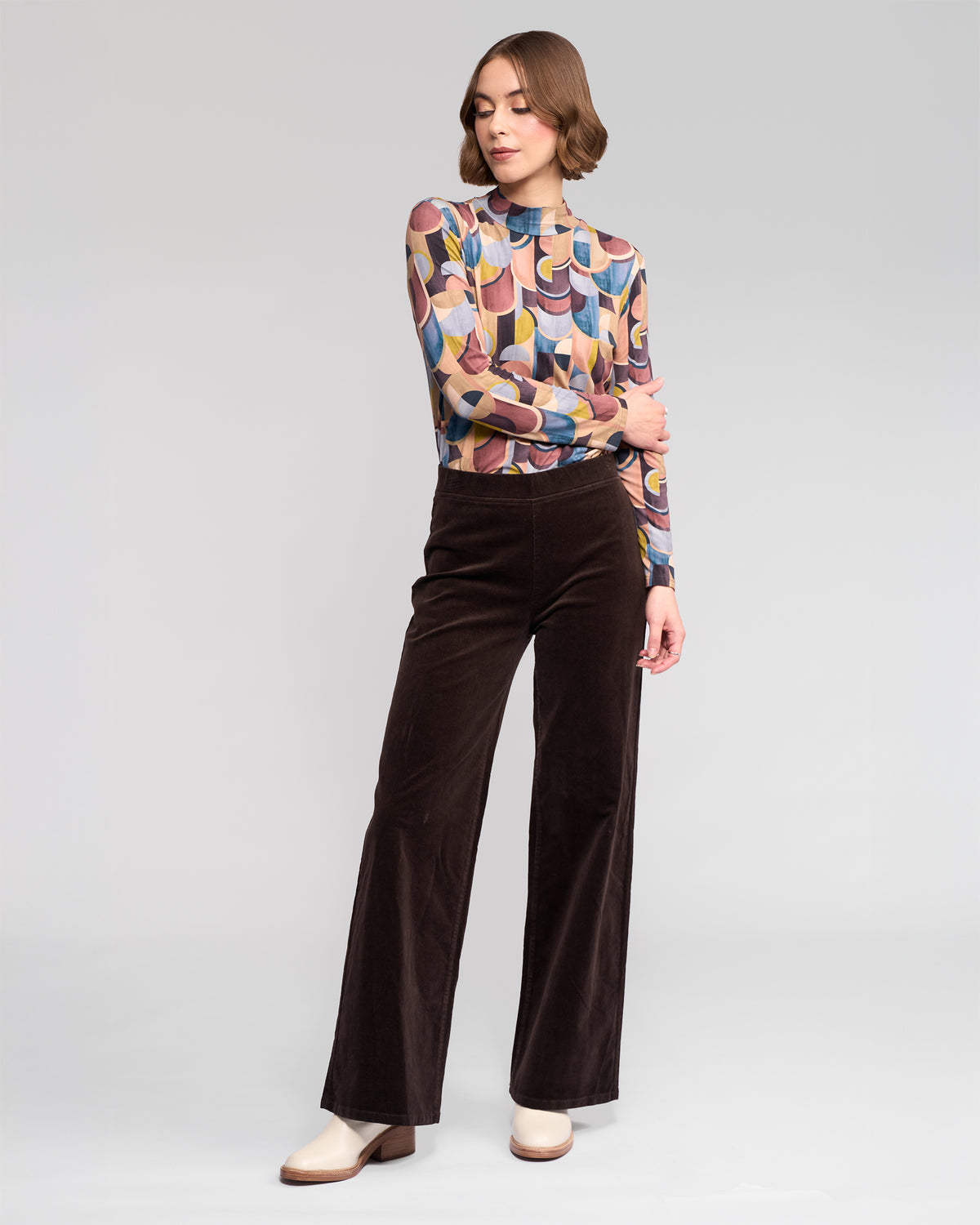 A person stands against a plain background wearing a colorful patterned long-sleeve top and Vassalli Loft Cord Pull On wide-leg pants in Walnut, paired with ivory ankle boots, with one arm resting on the opposite elbow.