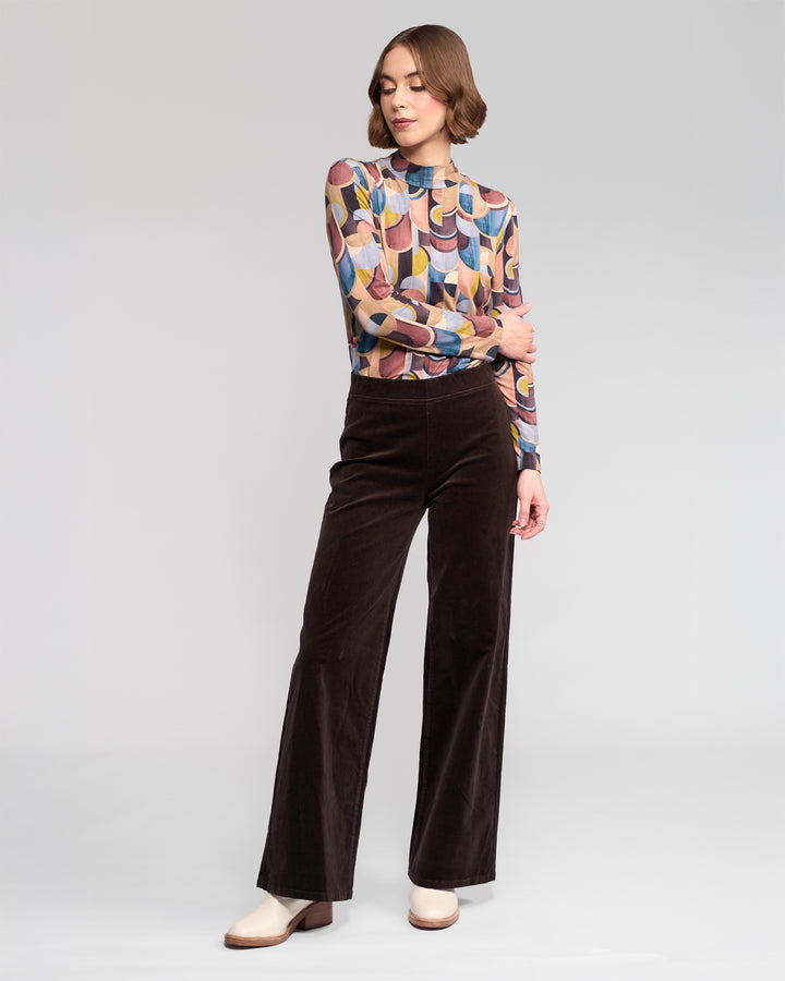 A person stands against a plain background wearing a colorful patterned long-sleeve top and Vassalli Loft Cord Pull On wide-leg pants in Walnut, paired with ivory ankle boots, with one arm resting on the opposite elbow.
