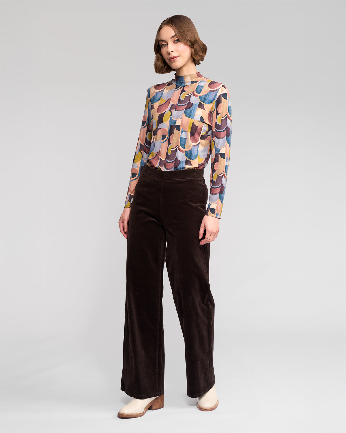 A woman with short wavy hair and a neutral expression stands against a plain background wearing a vibrant long-sleeve top, beige shoes, and Vassalli’s Loft Cord Pull On pants in Walnut—a dark brown wide-leg style with an elastic waistband.