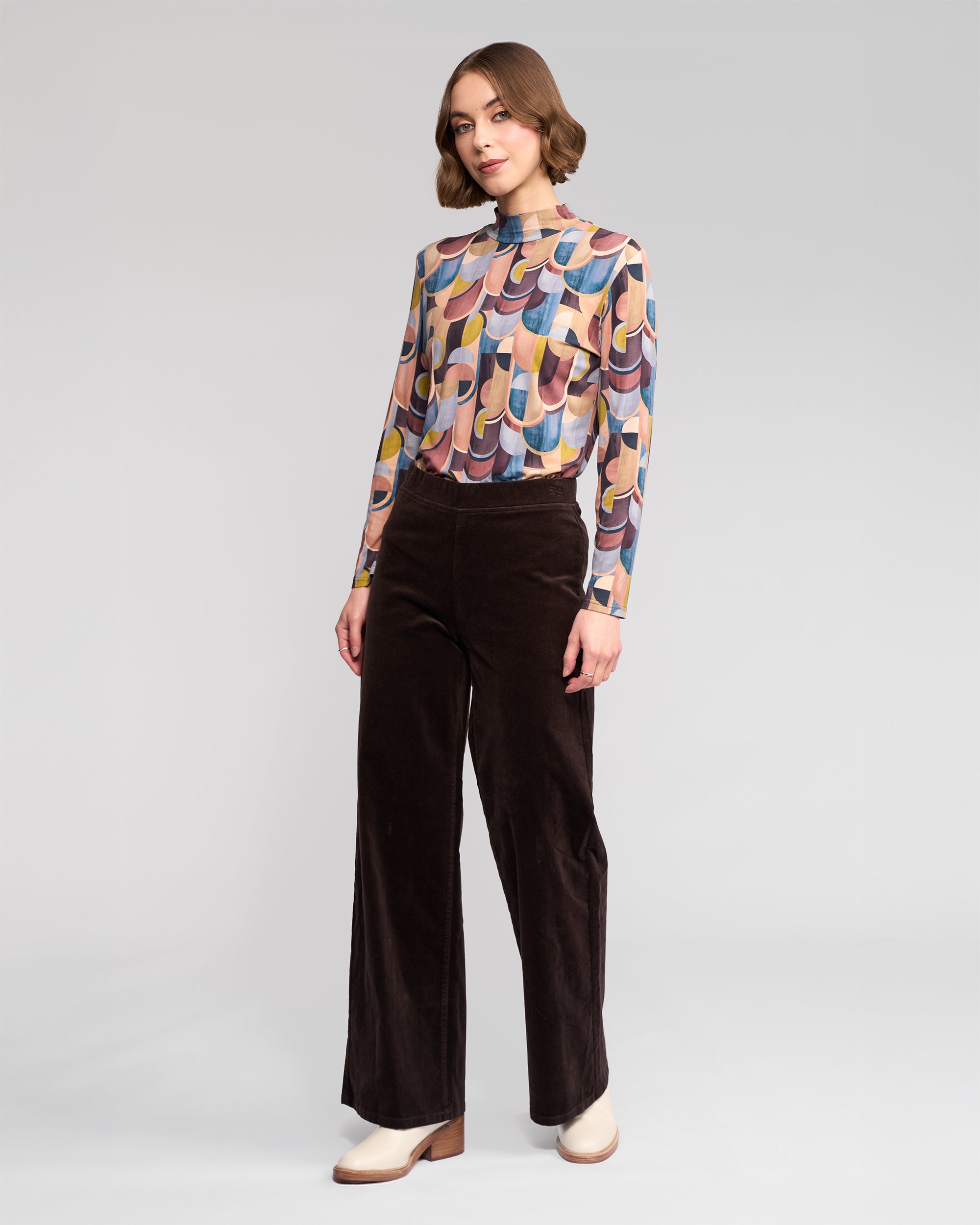 A woman with short wavy hair and a neutral expression stands against a plain background wearing a vibrant long-sleeve top, beige shoes, and Vassalli’s Loft Cord Pull On pants in Walnut—a dark brown wide-leg style with an elastic waistband.