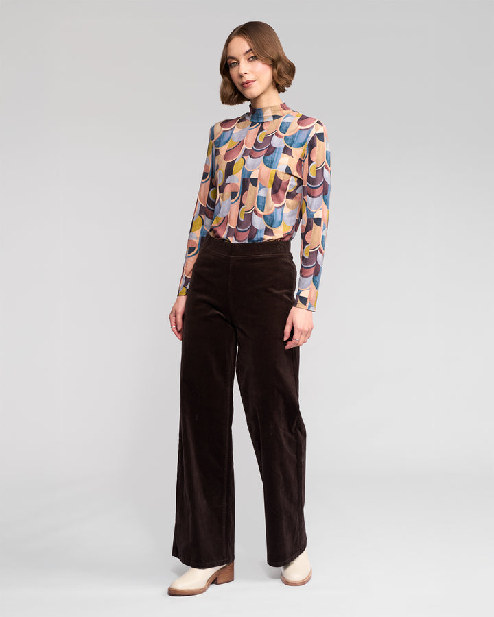 A woman with short wavy hair and a neutral expression stands against a plain background wearing a vibrant long-sleeve top, beige shoes, and Vassalli’s Loft Cord Pull On pants in Walnut—a dark brown wide-leg style with an elastic waistband.