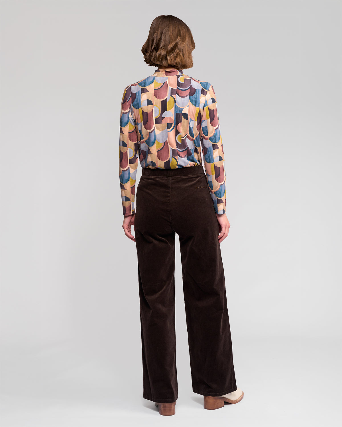 A person stands facing away, wearing Vassalli's Loft Cord Pull On pants in Walnut with an elasticated waistband, paired with a long-sleeve shirt featuring a colorful geometric pattern. The background is plain, light gray.