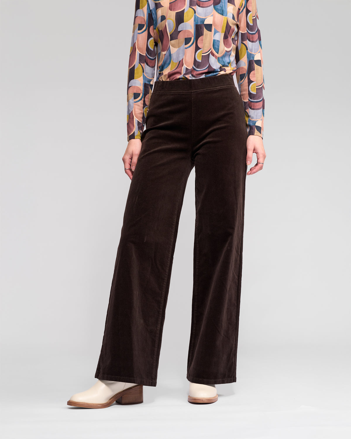 A person wears Vassalli Loft Cord Pull On - Walnut pants with an elasticated waistband, a colorful geometric-patterned long-sleeve shirt, and off-white block-heeled shoes against a plain background. Their face is not visible.