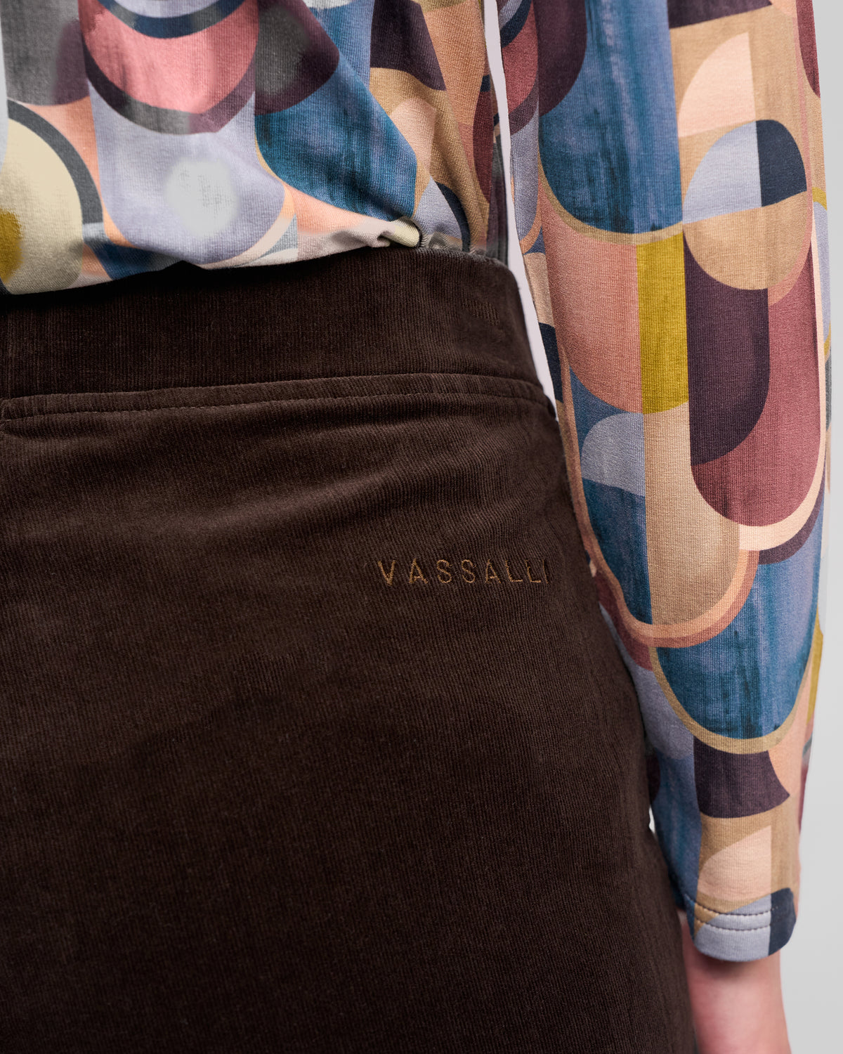 A close-up of a person wearing a colorful, geometric-patterned shirt and Vassalli Loft Cord Pull On - Walnut pants, with the word "VASSALLI" embroidered above the pocket.