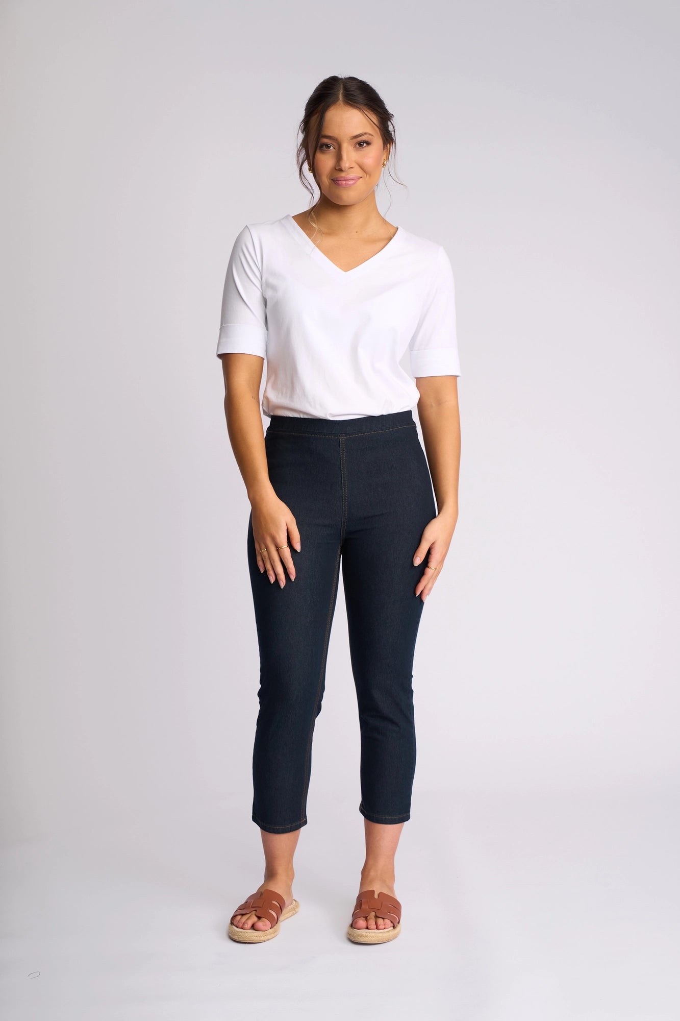 A woman stands facing forward against a light, plain background, wearing a white V-neck T-shirt, brown sandals, and the Vassalli Harbour 7/8 Pant in Indigo Denim (Contrast Stitch).