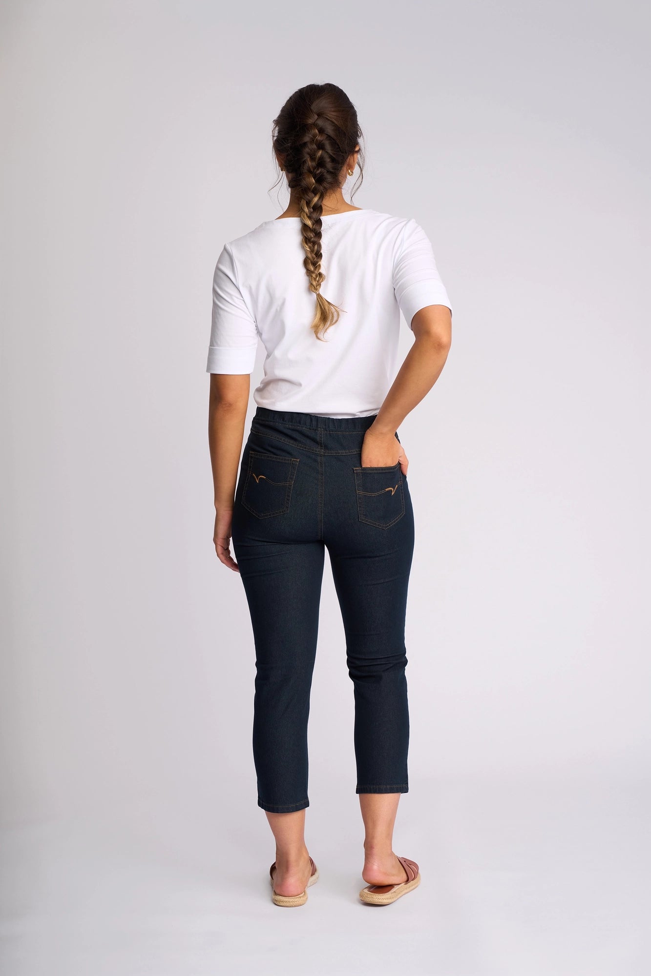 A woman stands facing away, wearing a white shirt, the Vassalli Harbour 7/8 Pant - Indigo Denim (Contrast Stitch), and sandals. She has a long braid down her back and one hand in her pocket. The background is plain and light-colored.