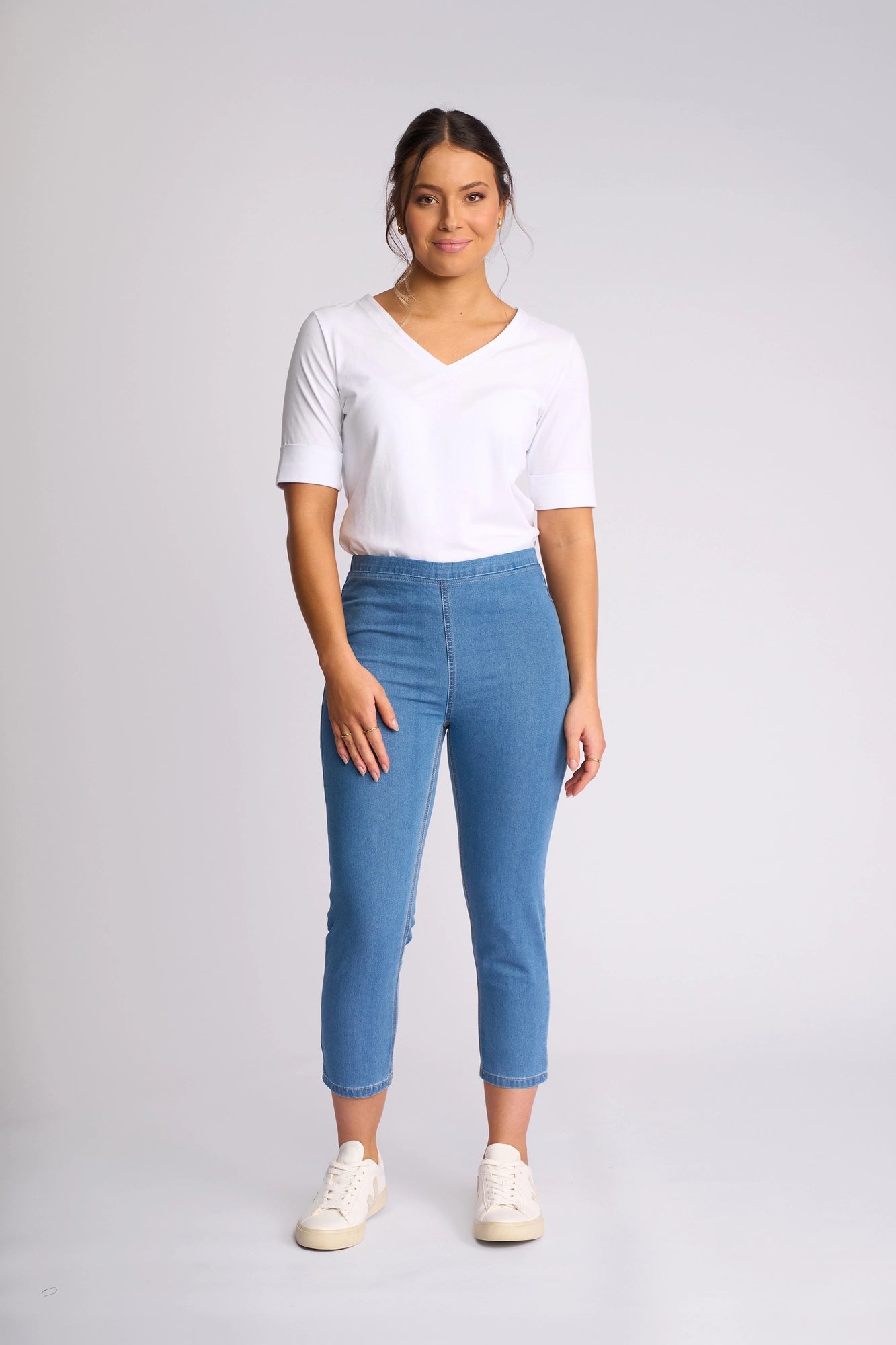 A woman stands facing forward against a plain light background, wearing a white short-sleeve top paired with Vassalli’s Harbour 7/8 Pant in Light Denim (Contrast Stitch)—high-waisted, cropped jeans crafted from stretch denim—and white sneakers.