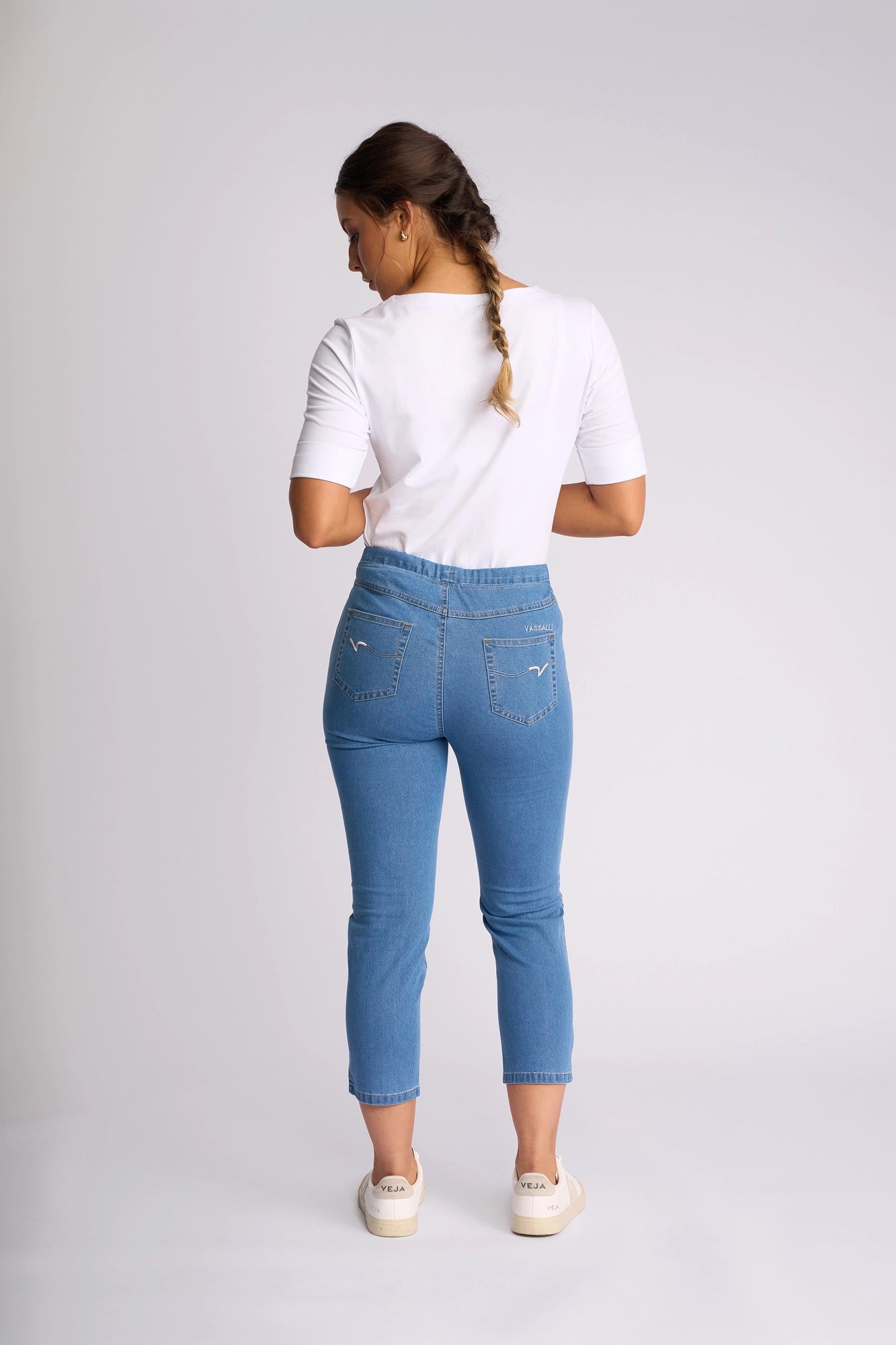 A woman stands facing away against a plain light background, her brown hair in a braid. She wears a white short-sleeve shirt, white sneakers, and the Vassalli Harbour 7/8 Pant - Light Denim (Contrast Stitch): high-waisted, skinny-leg pull-on jeans in stretch denim.
