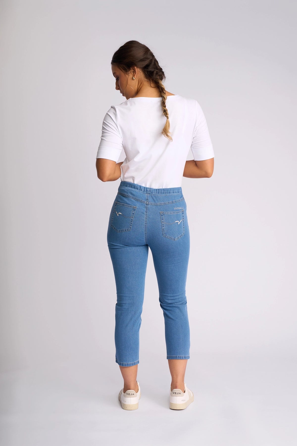 A woman stands facing away against a plain light background, her brown hair in a braid. She wears a white short-sleeve shirt, white sneakers, and the Vassalli Harbour 7/8 Pant - Light Denim (Contrast Stitch): high-waisted, skinny-leg pull-on jeans in stretch denim.