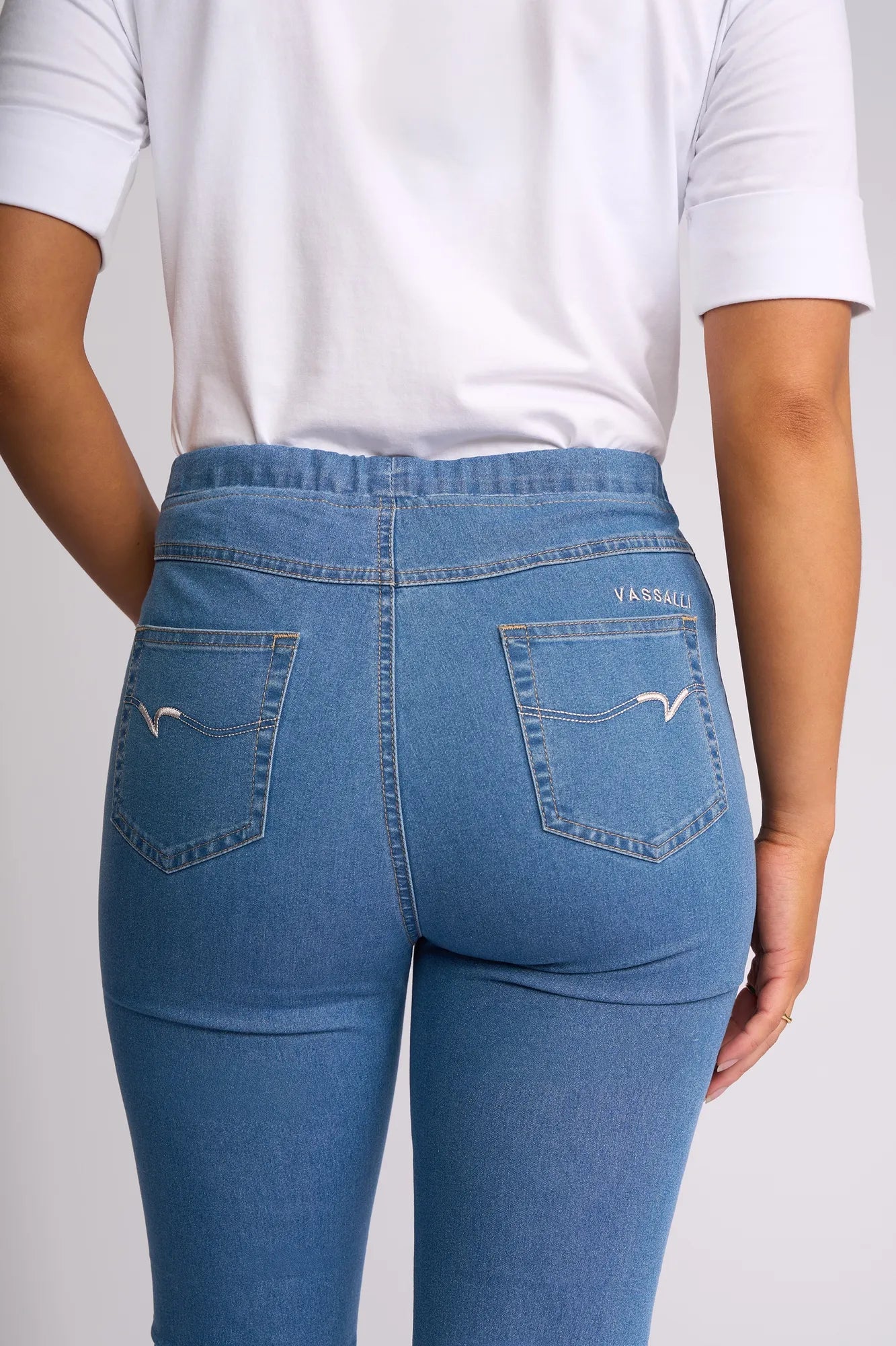 A person wearing a white shirt and blue Vassalli Harbour 7/8 Pant - Light Denim (Contrast Stitch) is shown from the back, focusing on the pants’ rear pockets and waistband. The stretch denim design by Vassalli combines comfort with contemporary style in a modern, 7/8 length silhouette.