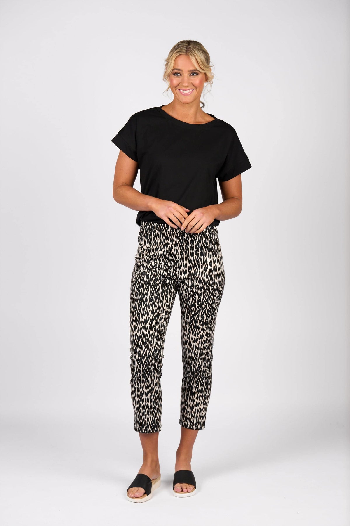 A woman smiles against a white background, wearing a black short-sleeve top, black slide sandals, and Vassalli's Harbour 7/8 Pant - Beau: a patterned black-and-white pull-on skinny leg pant in stretch fabric.