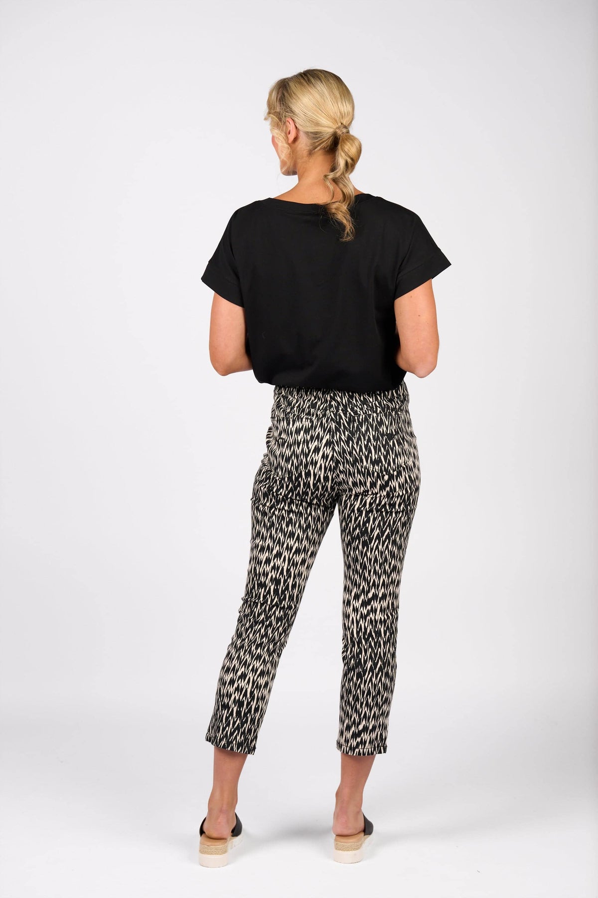 A woman with a loose blonde braid wears Vassalli's Harbour 7/8 Pant - Beau, featuring a black and white pattern and skinny leg fit, paired with a black short-sleeve top and heeled sandals against a plain white background.