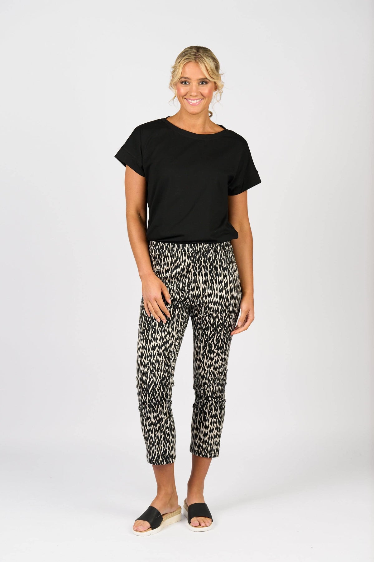 A woman stands smiling against a white background, wearing the Vassalli Harbour 7/8 Pant - Beau in a black-and-white pattern, paired with a black short-sleeve top and black slide sandals.
