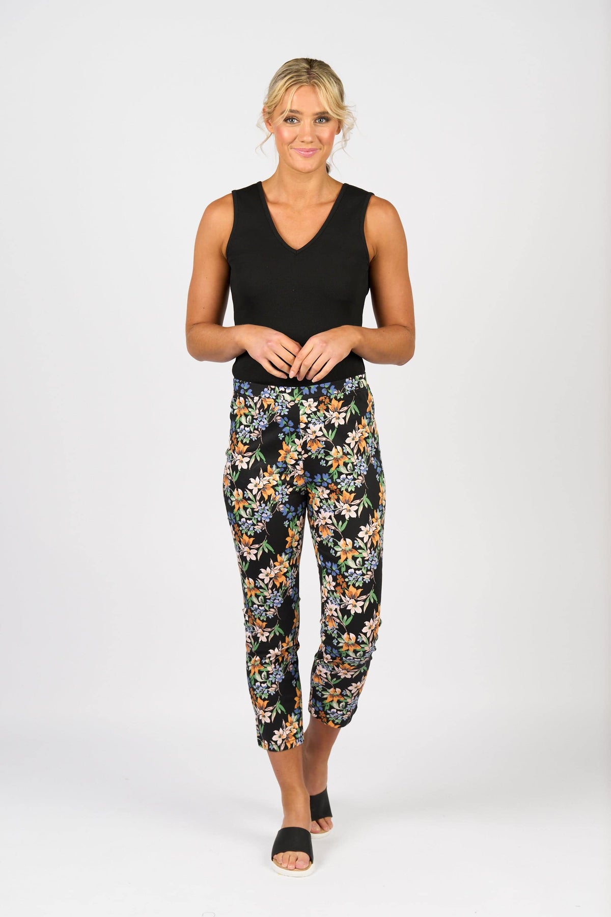 A woman with blonde hair in a loose updo wears the Vassalli Harbour 7/8 Pant - Grove against a white background, styled with a sleeveless black top and black slide sandals. She smiles slightly, hands together.
