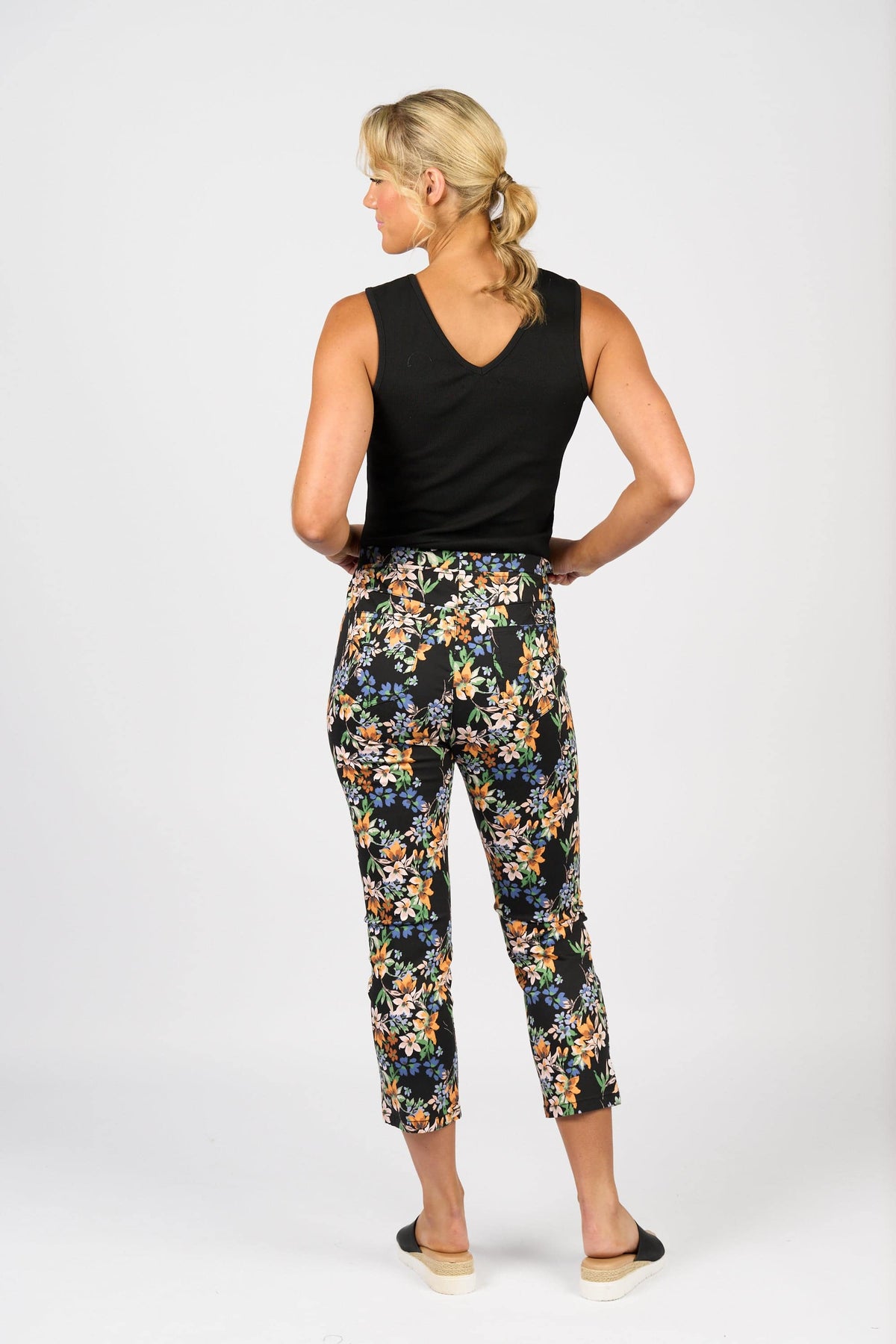 A woman with blonde hair in a ponytail wears the Vassalli Harbour 7/8 Pant - Grove and a black sleeveless top with black flats, standing facing away from the camera against a plain white background.