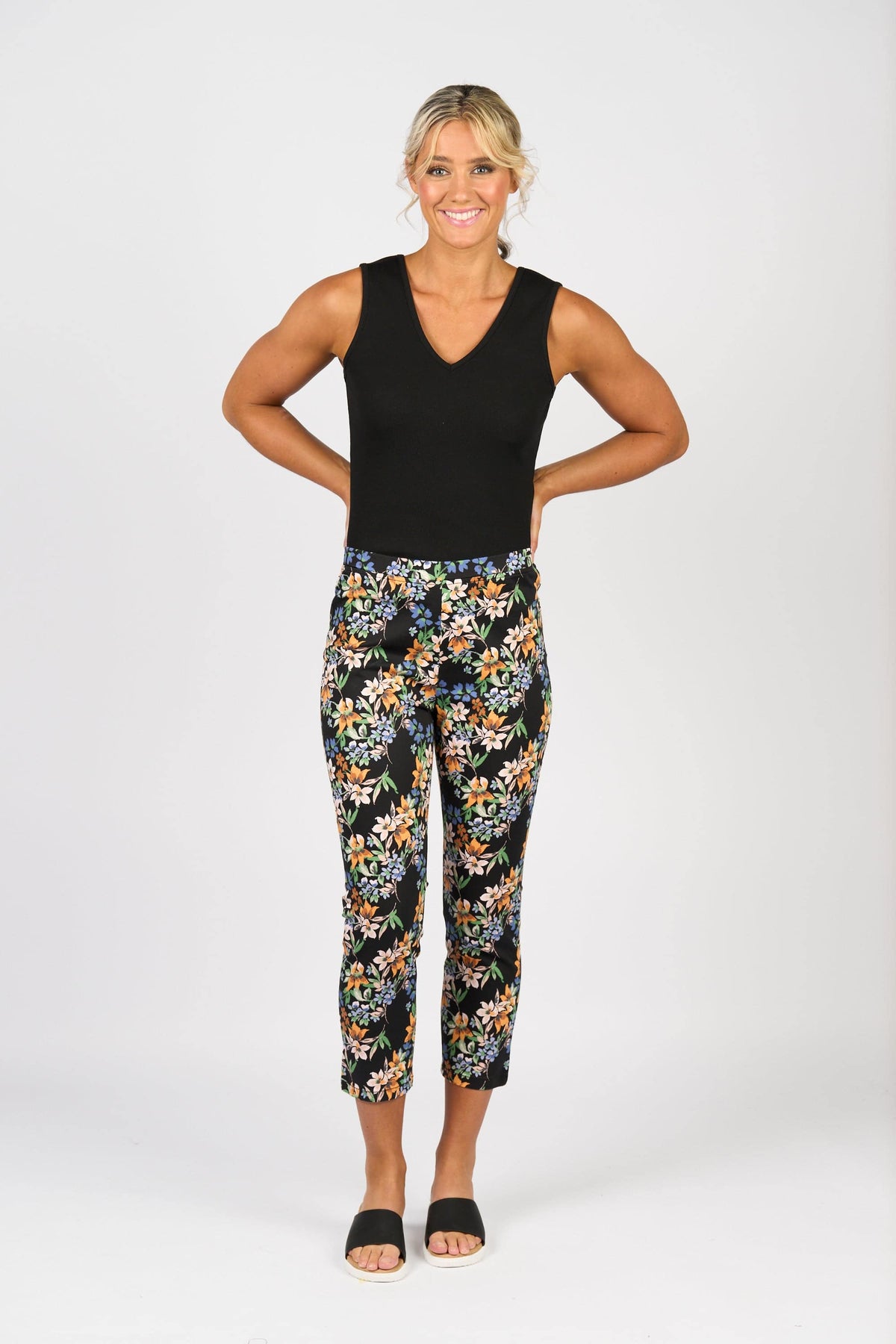 A smiling woman with blonde hair in a low bun poses against a white background, wearing a sleeveless black top and Vassalli Harbour 7/8 Pant - Grove, featuring stretch fabric, paired with black slide sandals. Her hands rest on her hips.