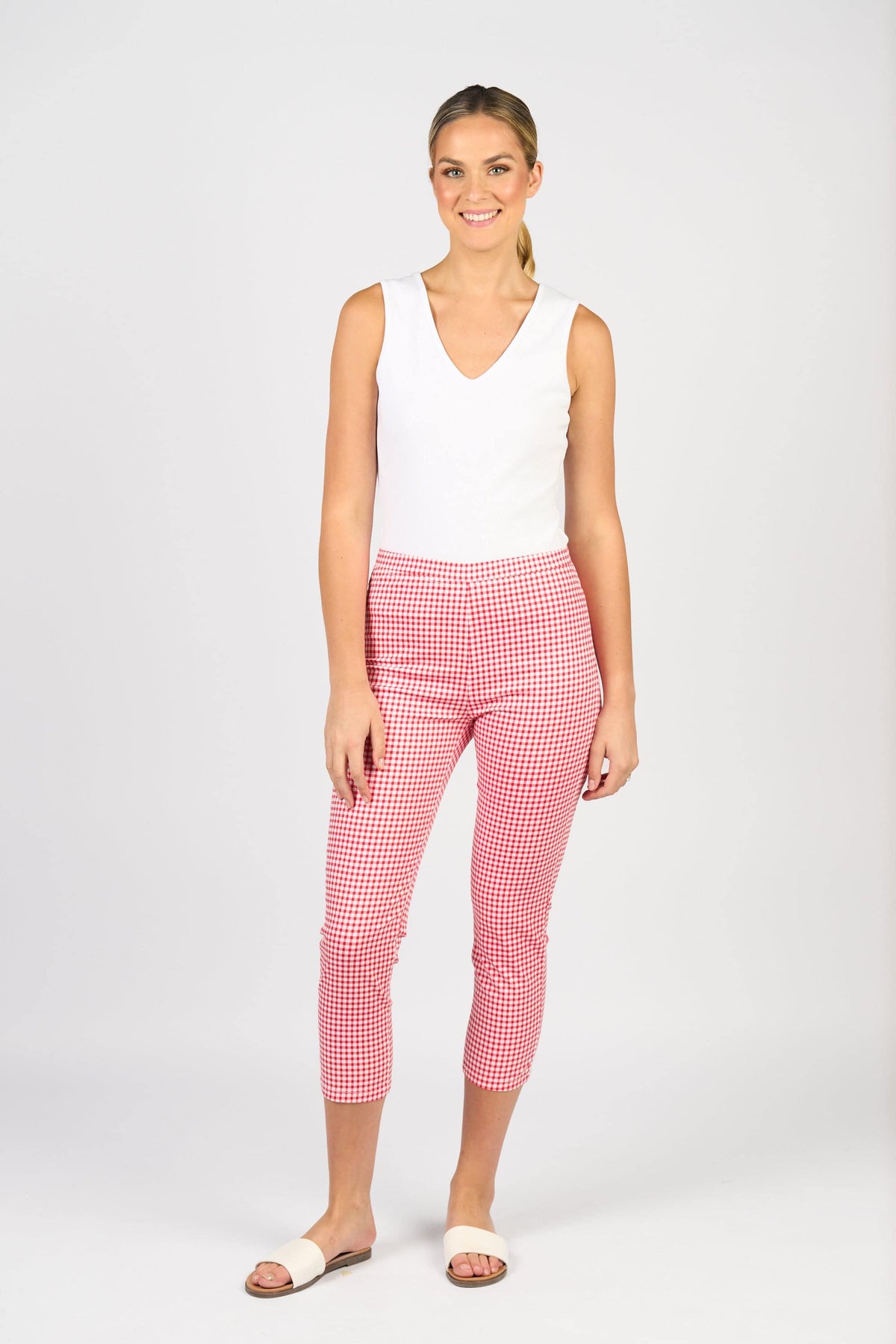 A woman smiles wearing the Vassalli Harbour 7/8 Pant in Red Gingham, featuring a skinny leg fit, paired with a sleeveless white top and white slide sandals against a plain light background.