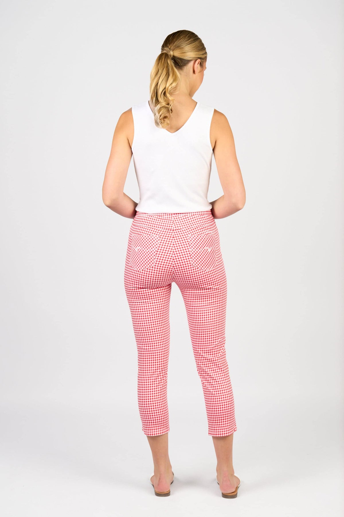 A woman with blonde hair in a ponytail faces away, wearing a white sleeveless top and Vassalli's Harbour 7/8 Pant in red gingham stretch fabric. She is barefoot against a plain white background.