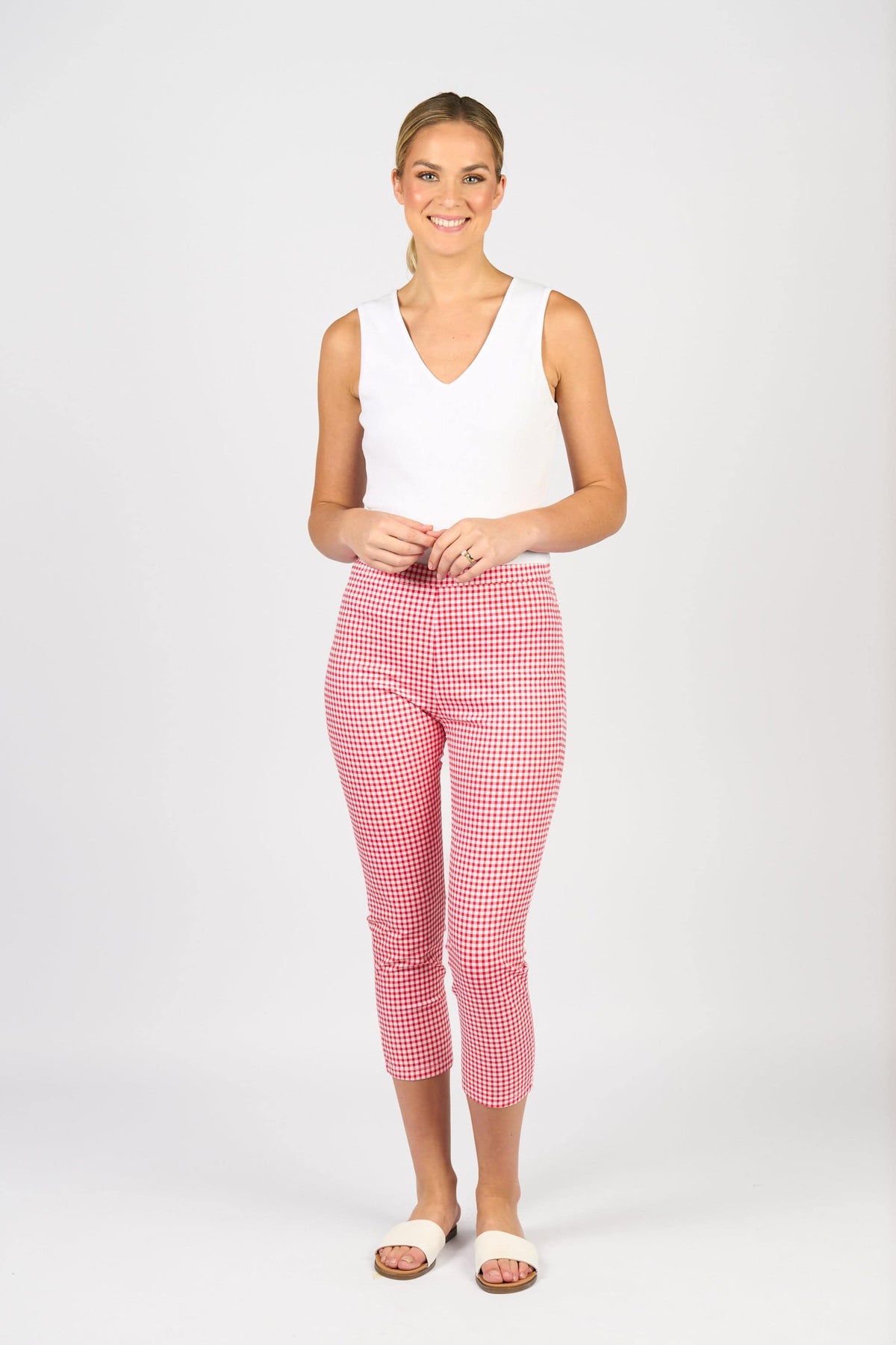 A woman smiles against a plain background, wearing a white sleeveless top, Vassalli Harbour 7/8 Pant in Red Gingham made from stretch fabric, and white slide sandals.