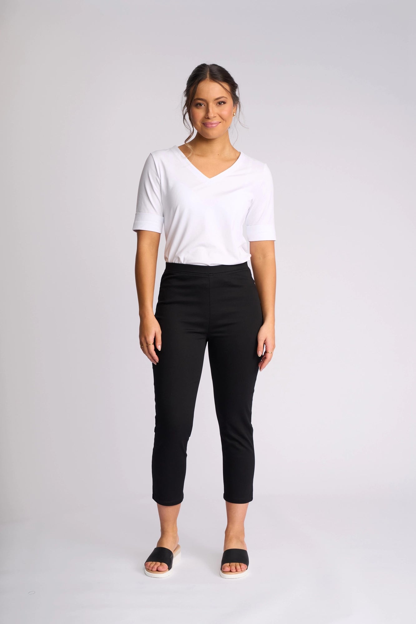 A woman stands and smiles against a plain light gray background, wearing a white V-neck T-shirt, black Vassalli Harbour 7/8 Pant crafted from stretch fabric in a skinny-leg pull-on style, and black open-toe sandals.