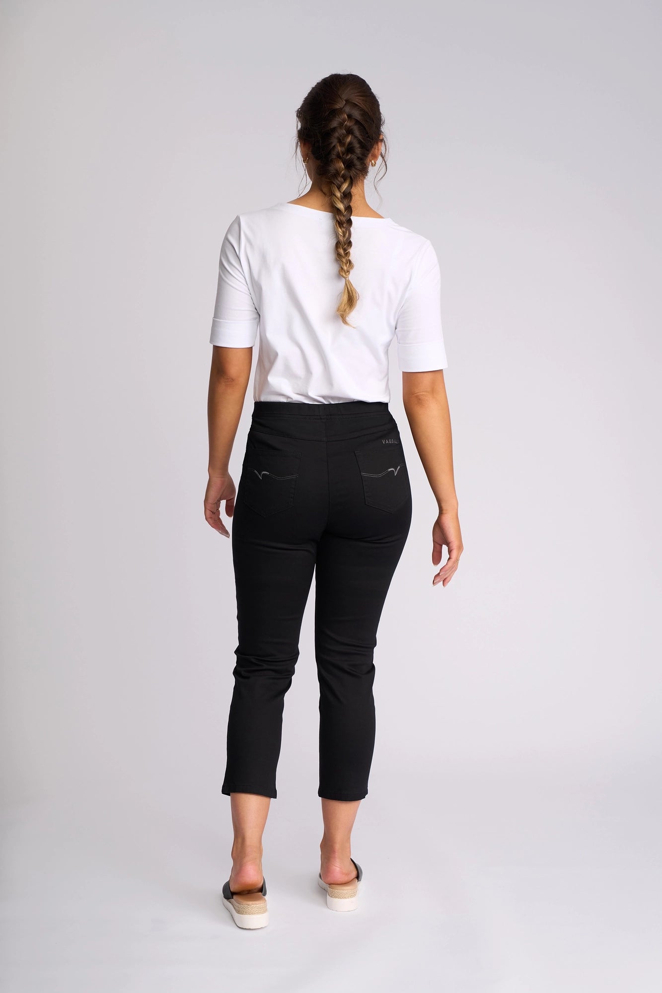 A woman with a braid stands facing away, wearing a white short-sleeve top and the Vassalli Harbour 7/8 Pant in Black—skinny-leg pull-on pants made from stretch fabric—paired with slip-on shoes against a plain light background.