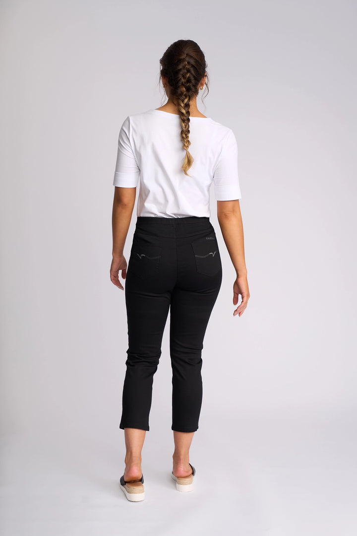 A woman with a braid stands facing away, wearing a white short-sleeve top and the Vassalli Harbour 7/8 Pant in Black—skinny-leg pull-on pants made from stretch fabric—paired with slip-on shoes against a plain light background.