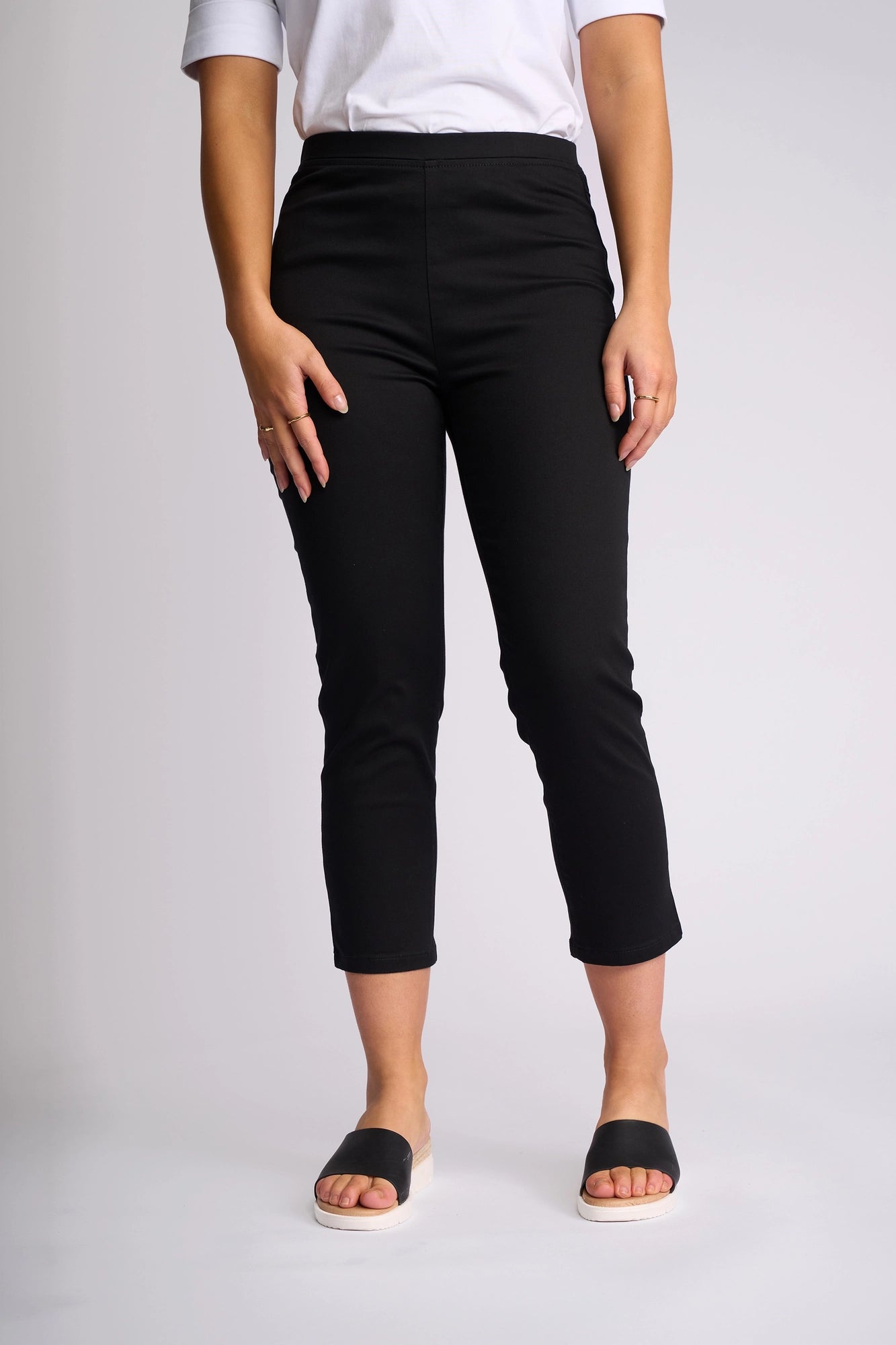 A person is shown standing against a plain light background, wearing the Vassalli Harbour 7/8 Pant in black—a skinny-leg, pull-on style crafted from stretch fabric—paired with a white short-sleeve top and black slide sandals. Their hands rest by their sides, and their face is not visible.
