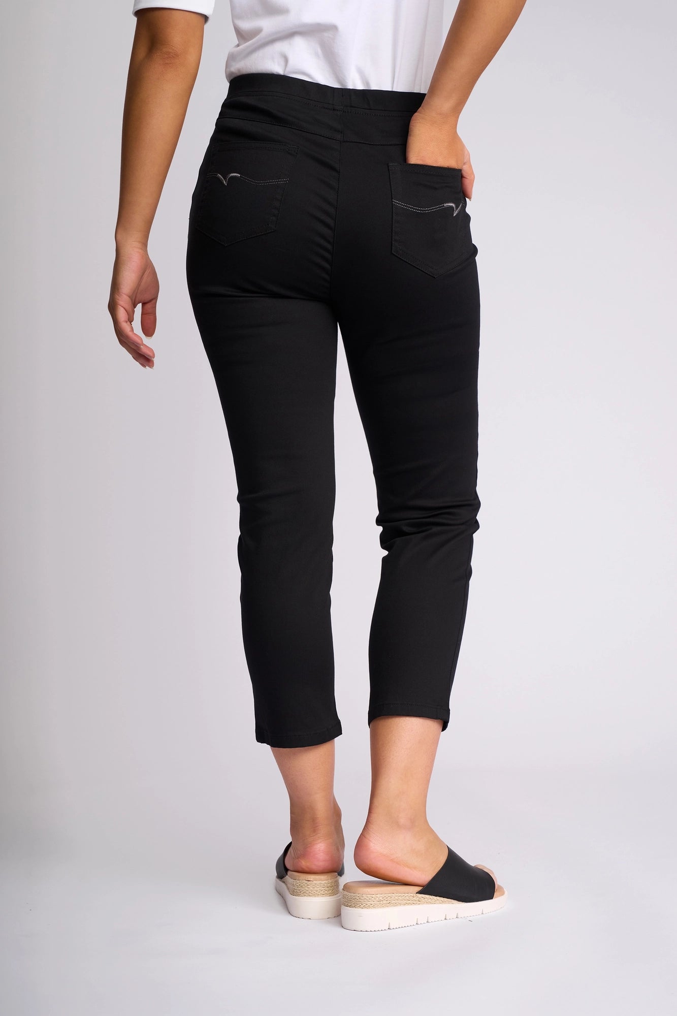 A person is seen from the back wearing Vassalli’s Harbour 7/8 Pant in black, featuring a skinny-leg pull-on design crafted from stretch fabric, paired with black slip-on sandals.