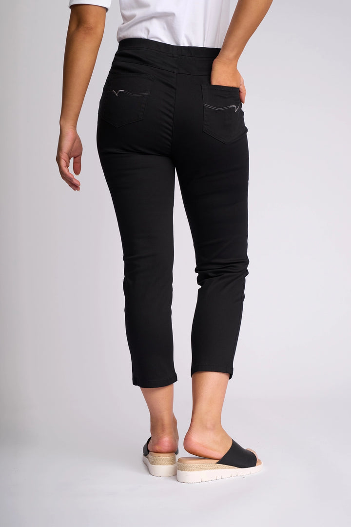 A person is seen from the back wearing Vassalli’s Harbour 7/8 Pant in black, featuring a skinny-leg pull-on design crafted from stretch fabric, paired with black slip-on sandals.