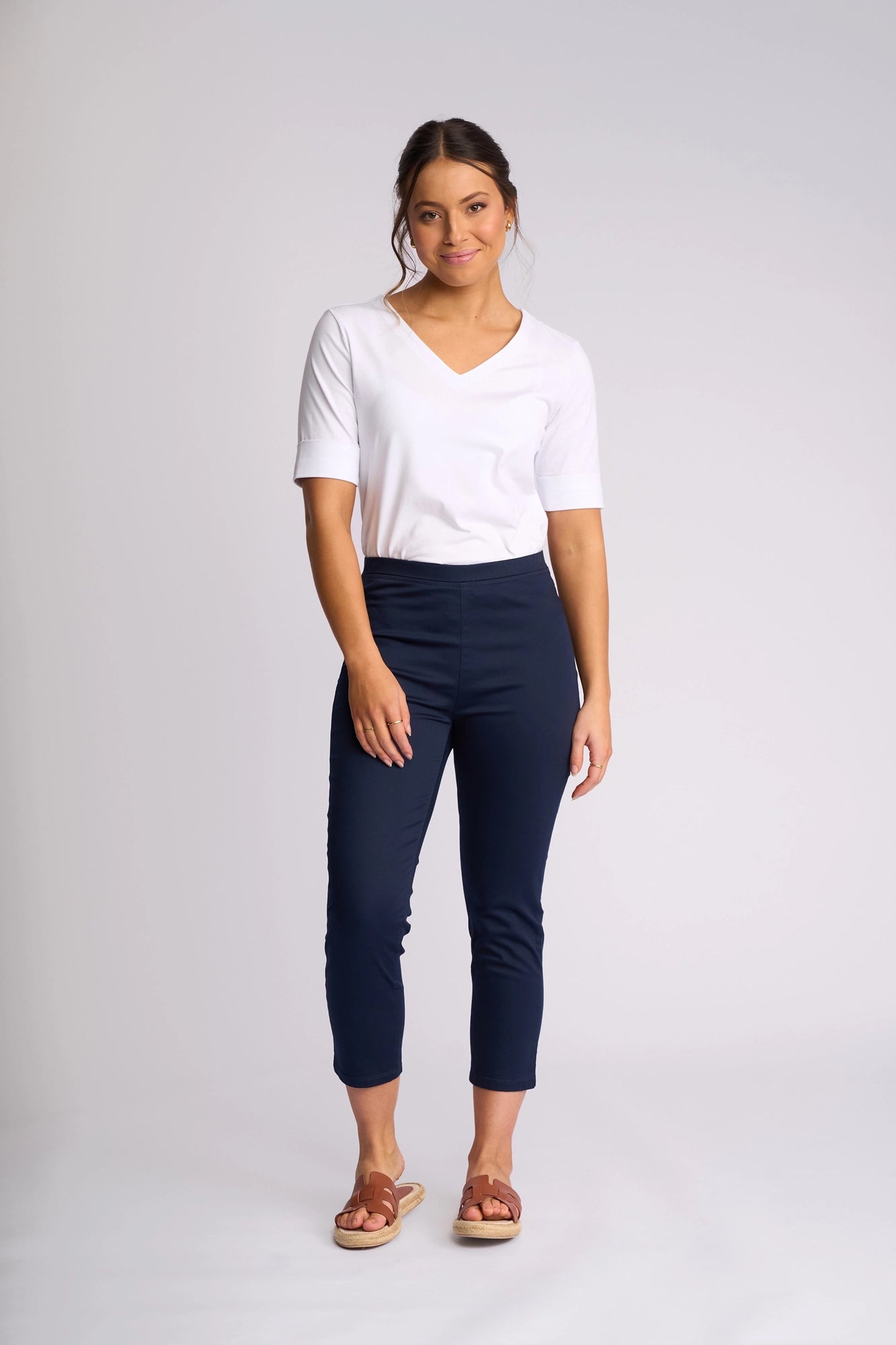A woman stands and smiles, wearing the Vassalli Harbour 7/8 Pant in Navy—a skinny-leg pull-on style crafted from stretch fabric—paired with a white short-sleeve shirt and brown sandals, set against a plain light background.